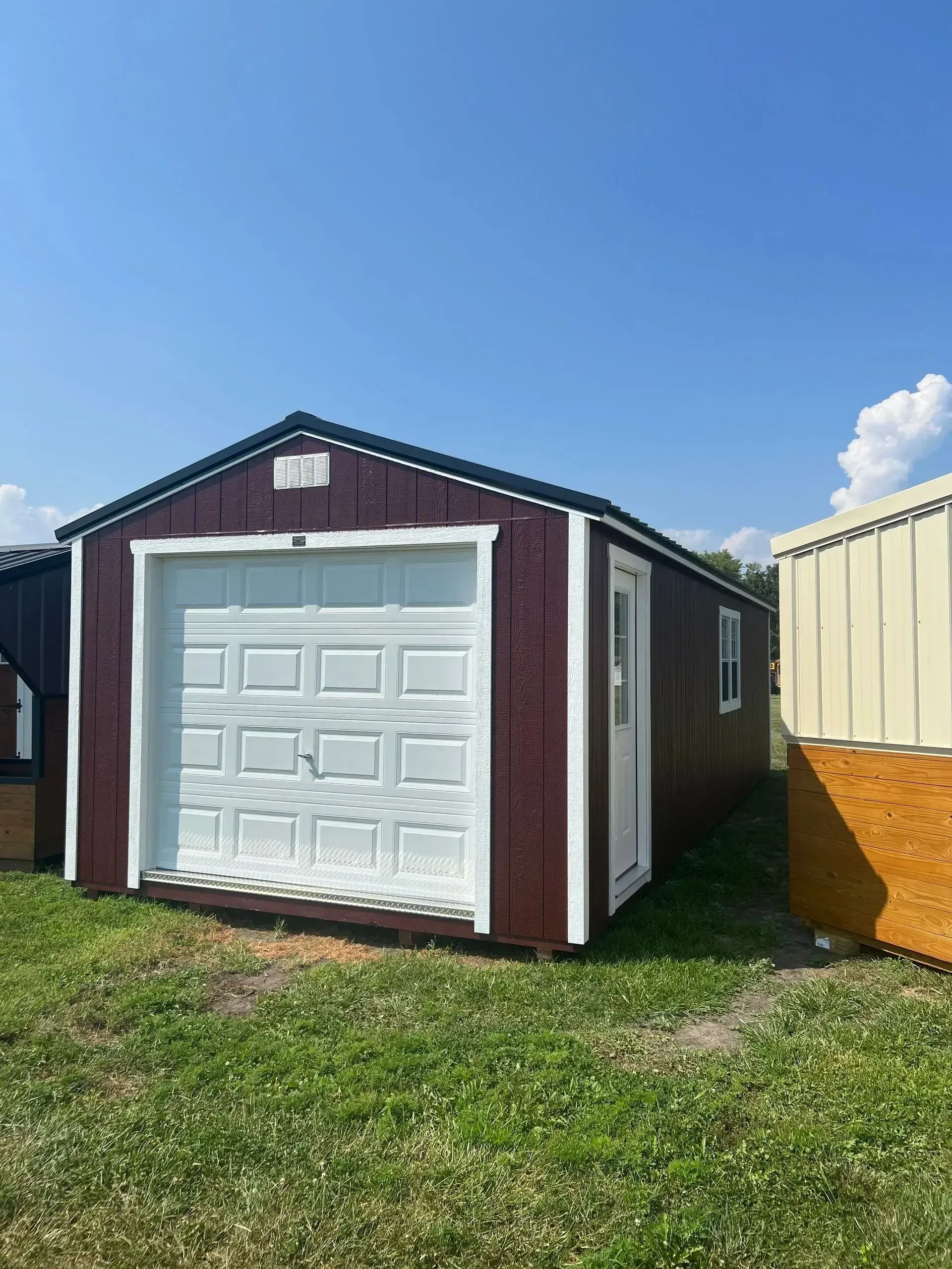 Brown shed with white garage door and trim, on green grass under blue sky.