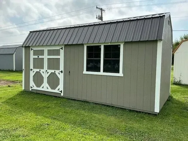 A tan shed with a dark gray roof, white trim, a door, and window sits on green grass.