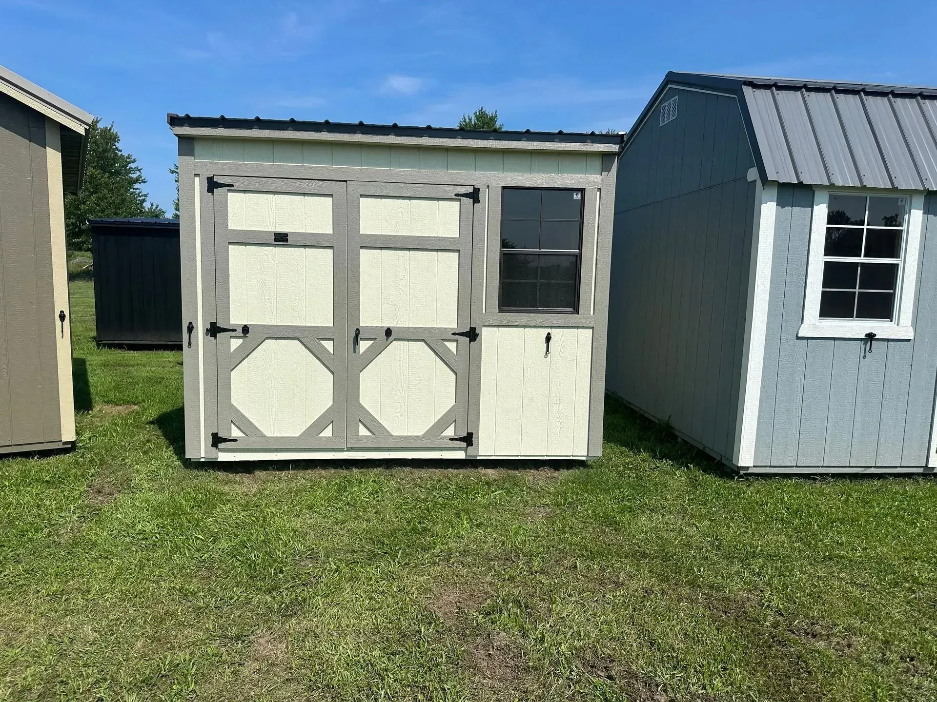 Tan shed with gray trim and double doors; sits on green grass with other sheds.