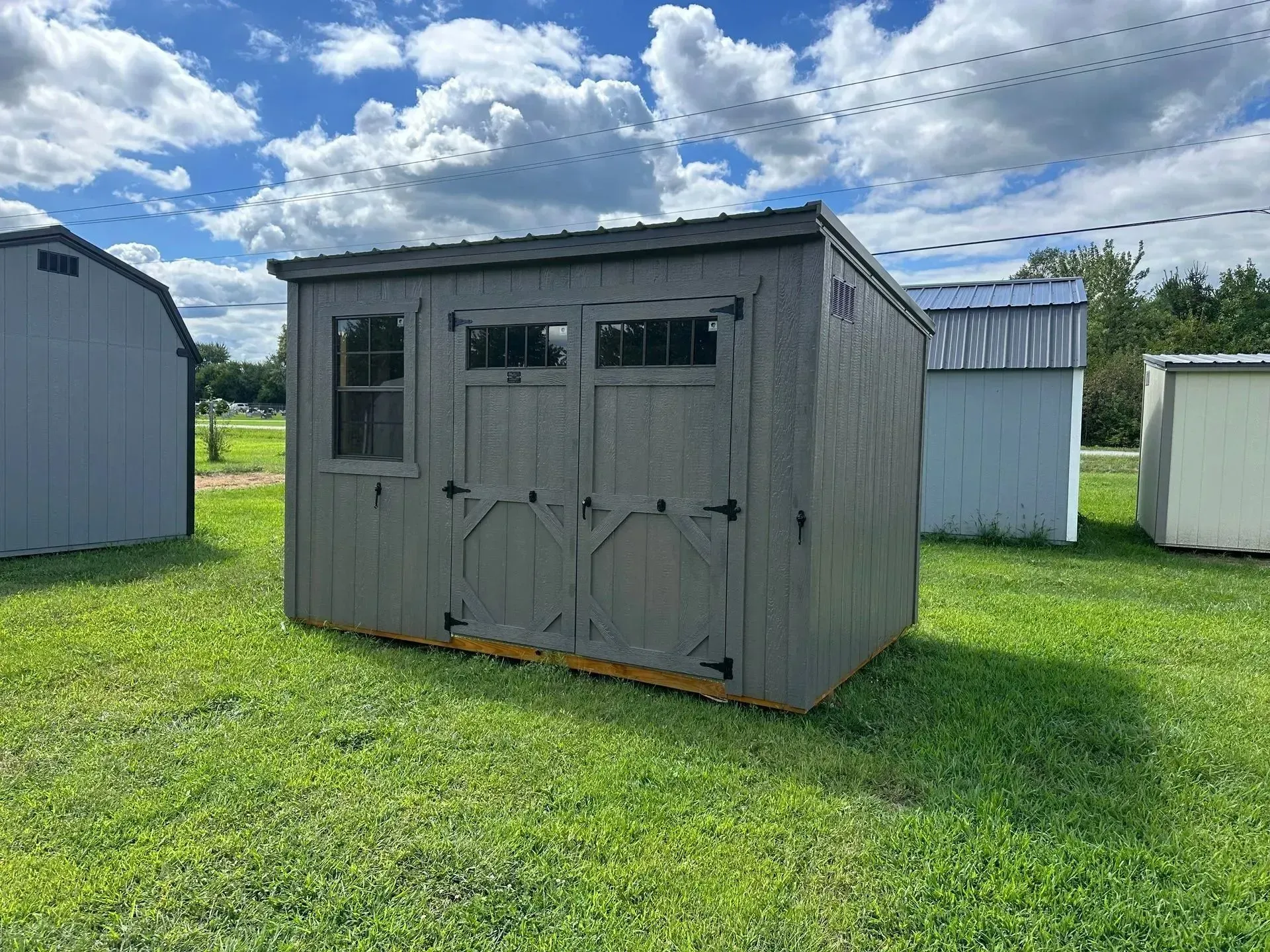Gray wooden shed with double doors and a window on a grassy field, under a blue sky.