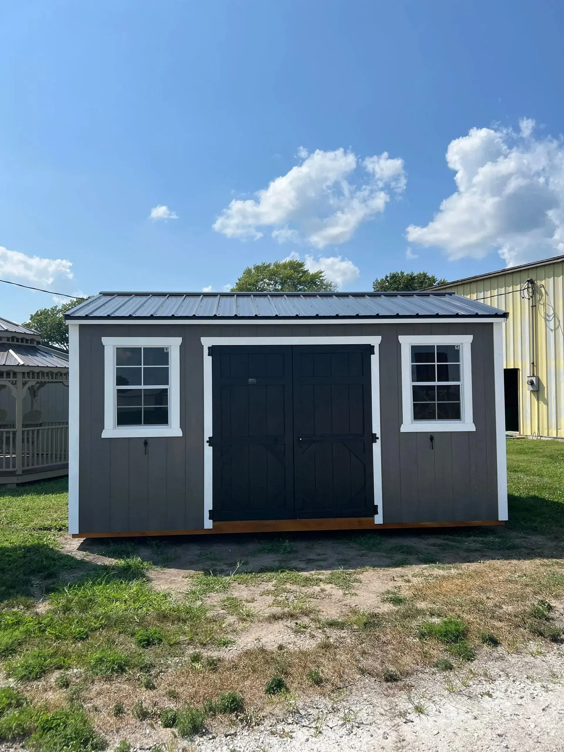 Gray storage shed with black doors, white-trimmed windows, and metal roof, on a grassy plot under a blue sky.
