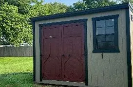 Tan shed with maroon double doors and a window, set in a grassy yard, surrounded by trees.