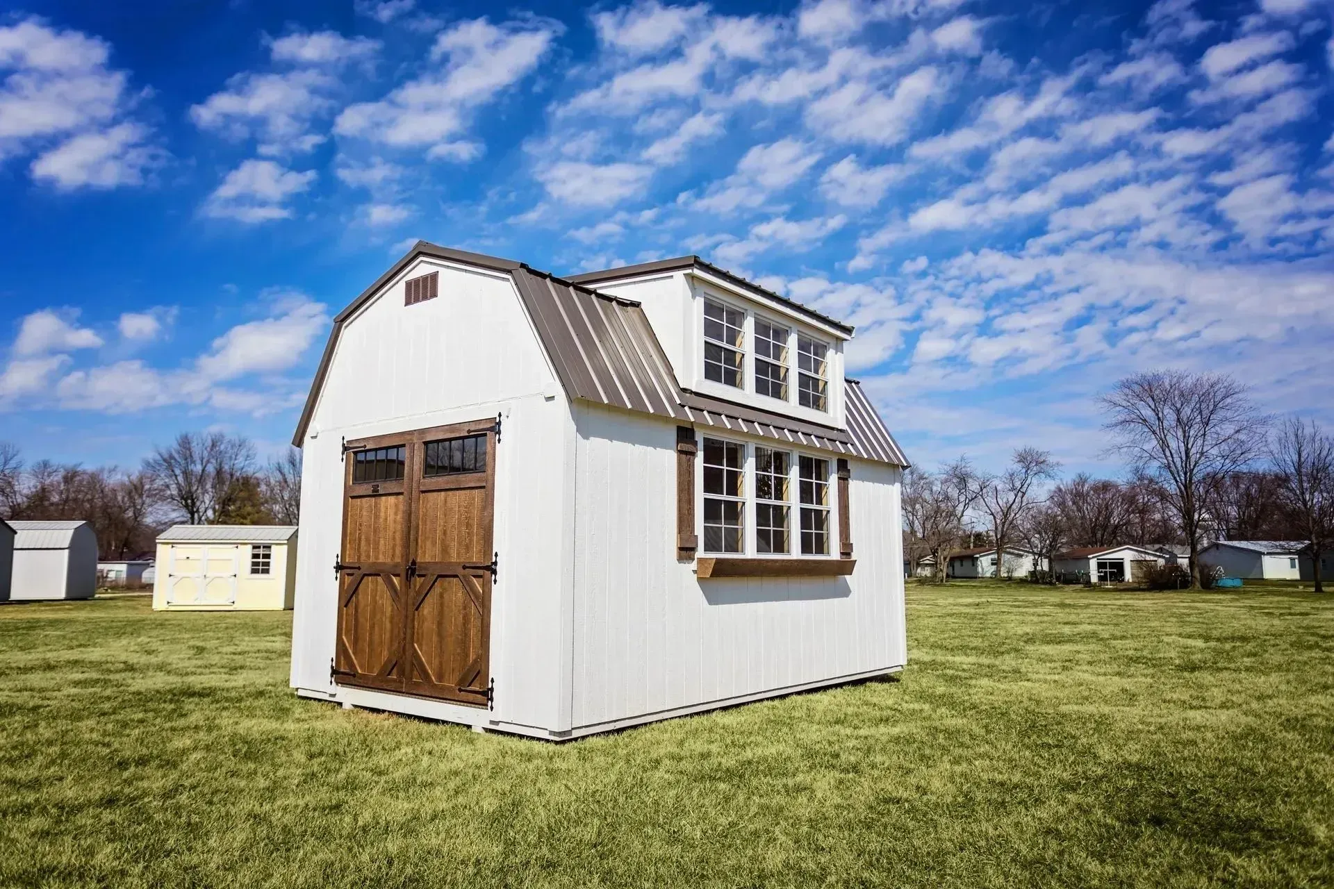 White barn-style shed with brown doors and metal roof, set in a grassy field under a blue sky.