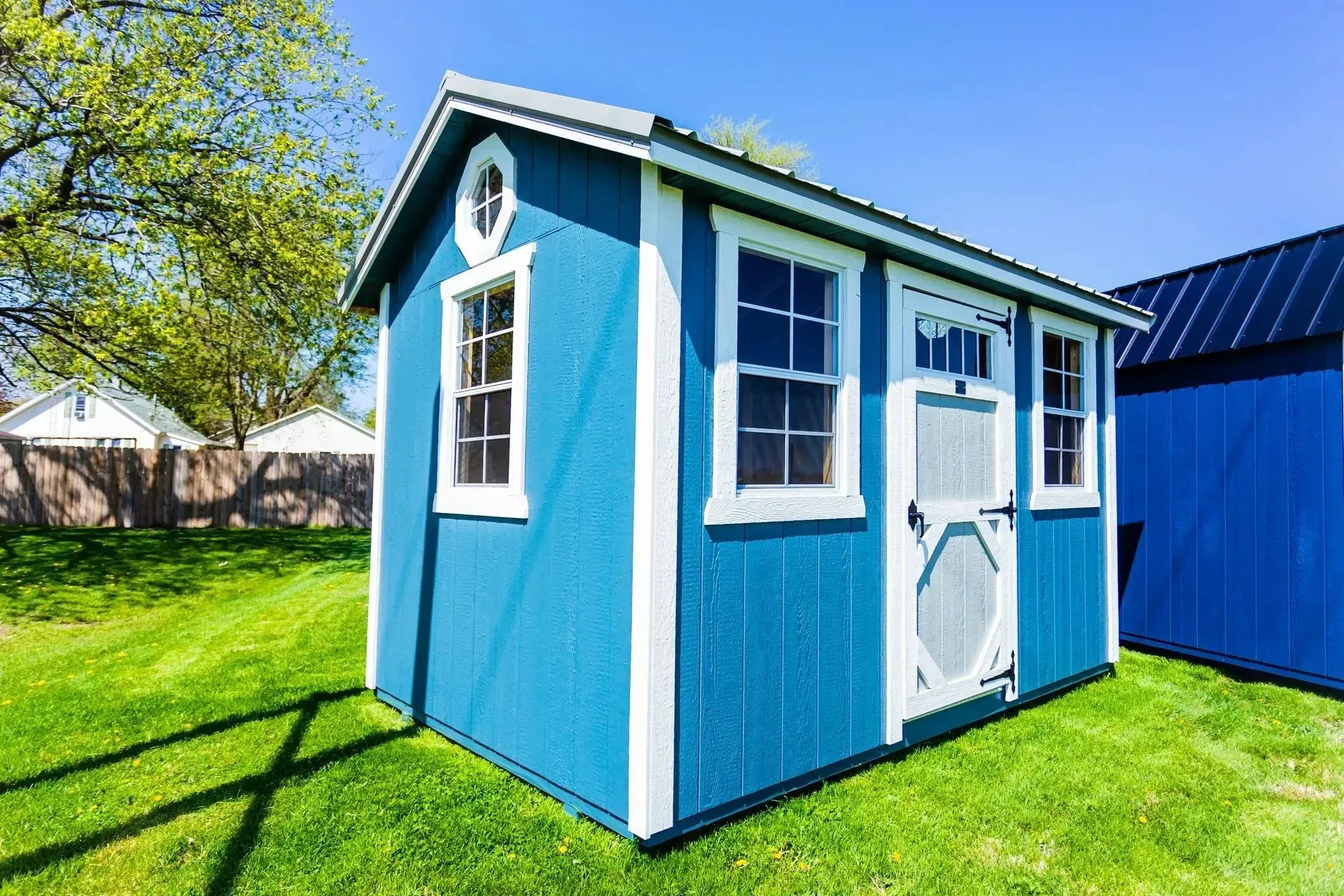 Blue shed with white trim, windows, and door, set on green grass under a blue sky.