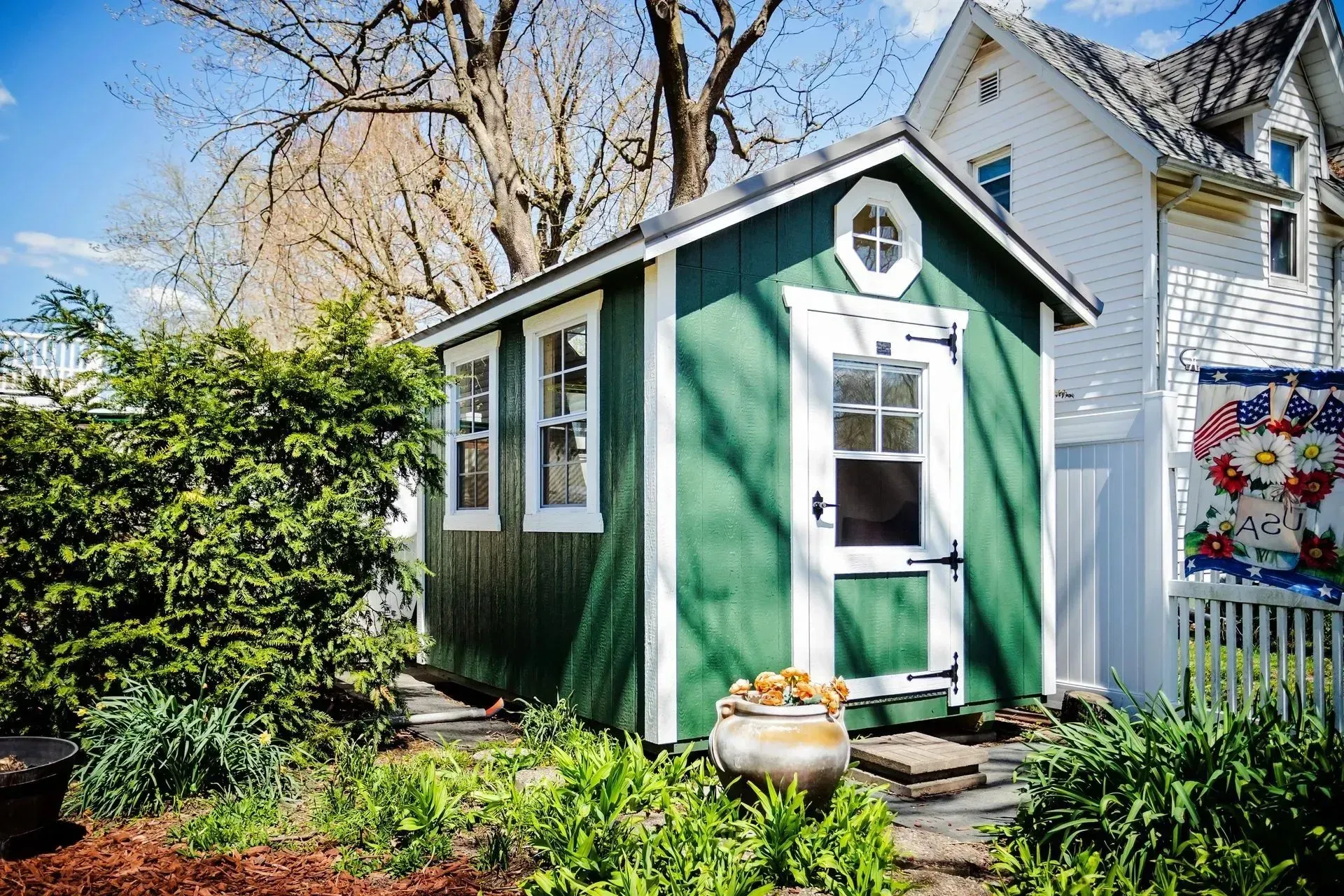 Green shed with white trim, a round window, and a white door in a garden.