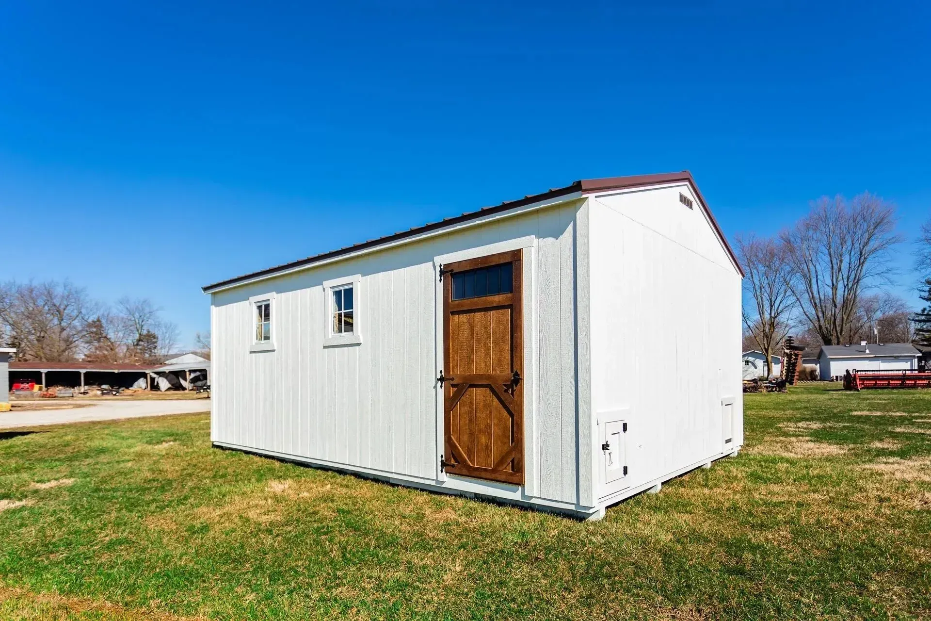 White shed with brown door and roof on green grass under blue sky.