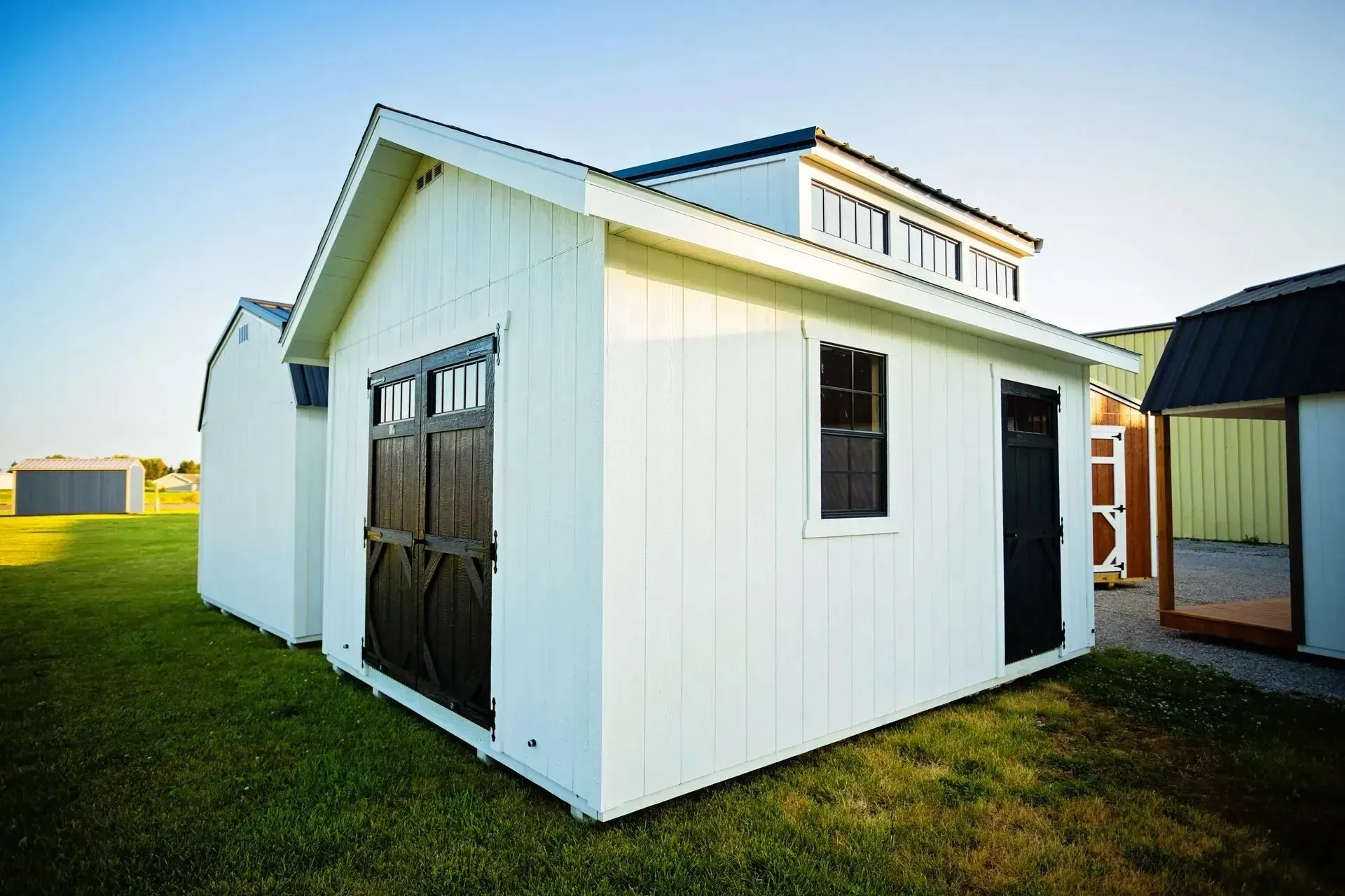 White shed with black doors and window on green grass, under a blue sky.