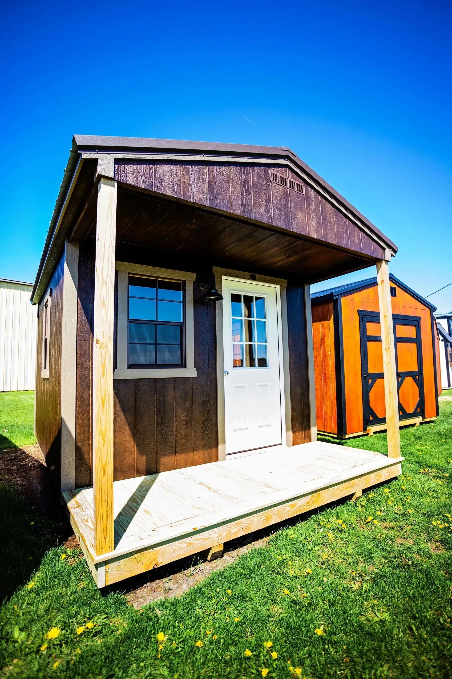 A small brown shed with a porch and white door, sitting on grass under a blue sky.