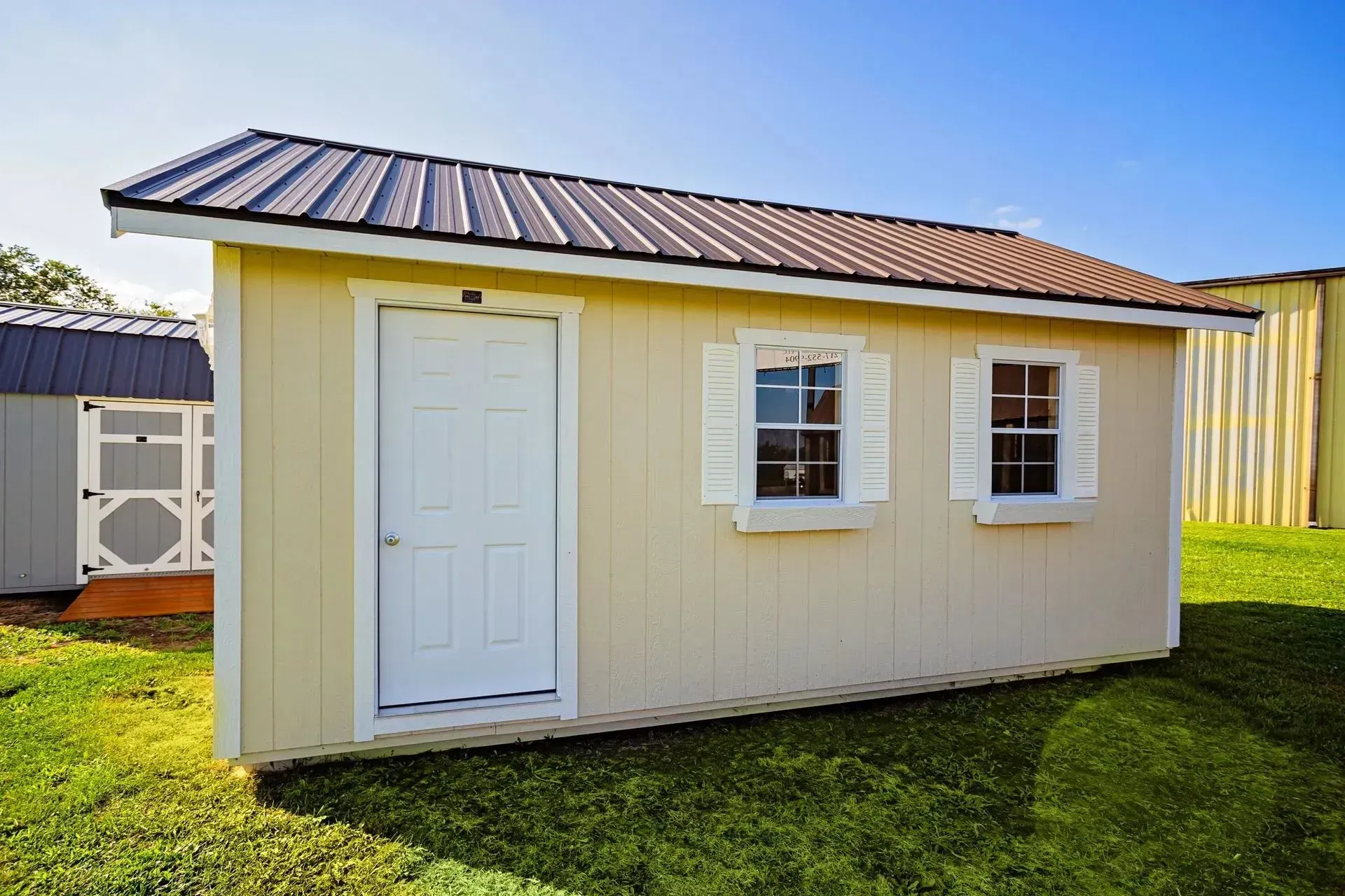 Tan storage shed with a brown metal roof, white door, and two windows with shutters.