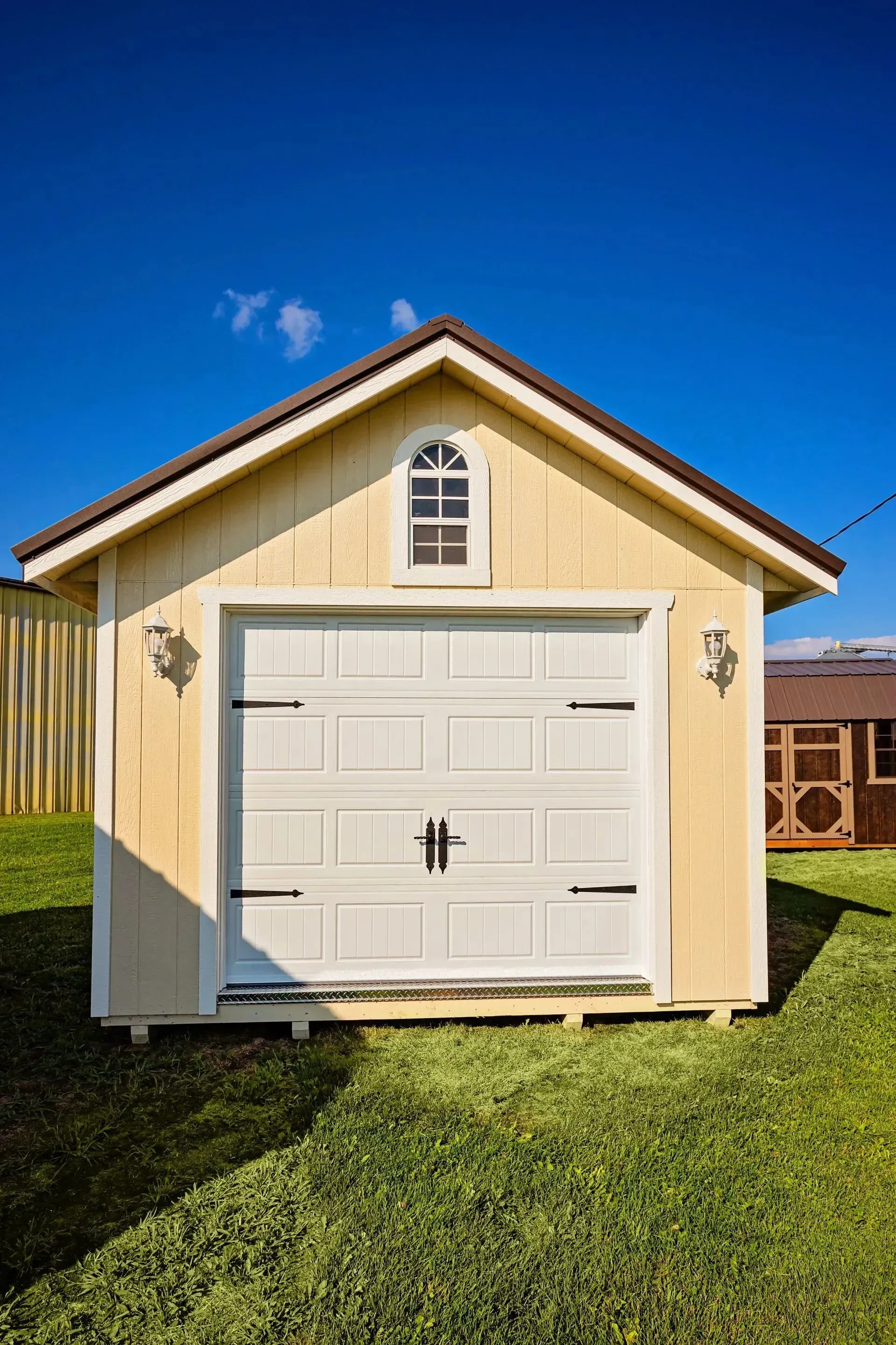 Yellow shed with white garage door, small arched window, on green grass, blue sky.