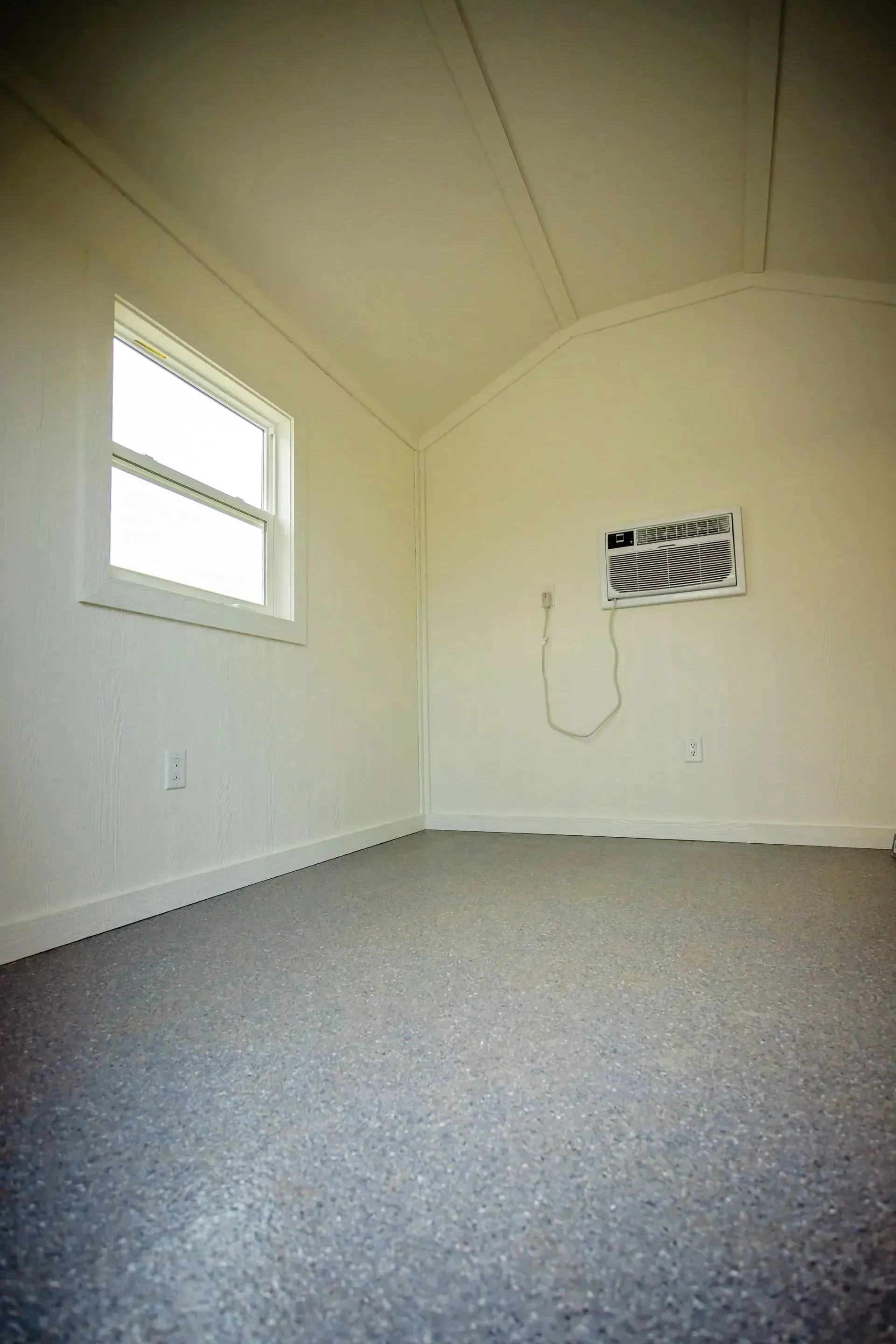 Empty white room with a window, air conditioner, and gray speckled floor.