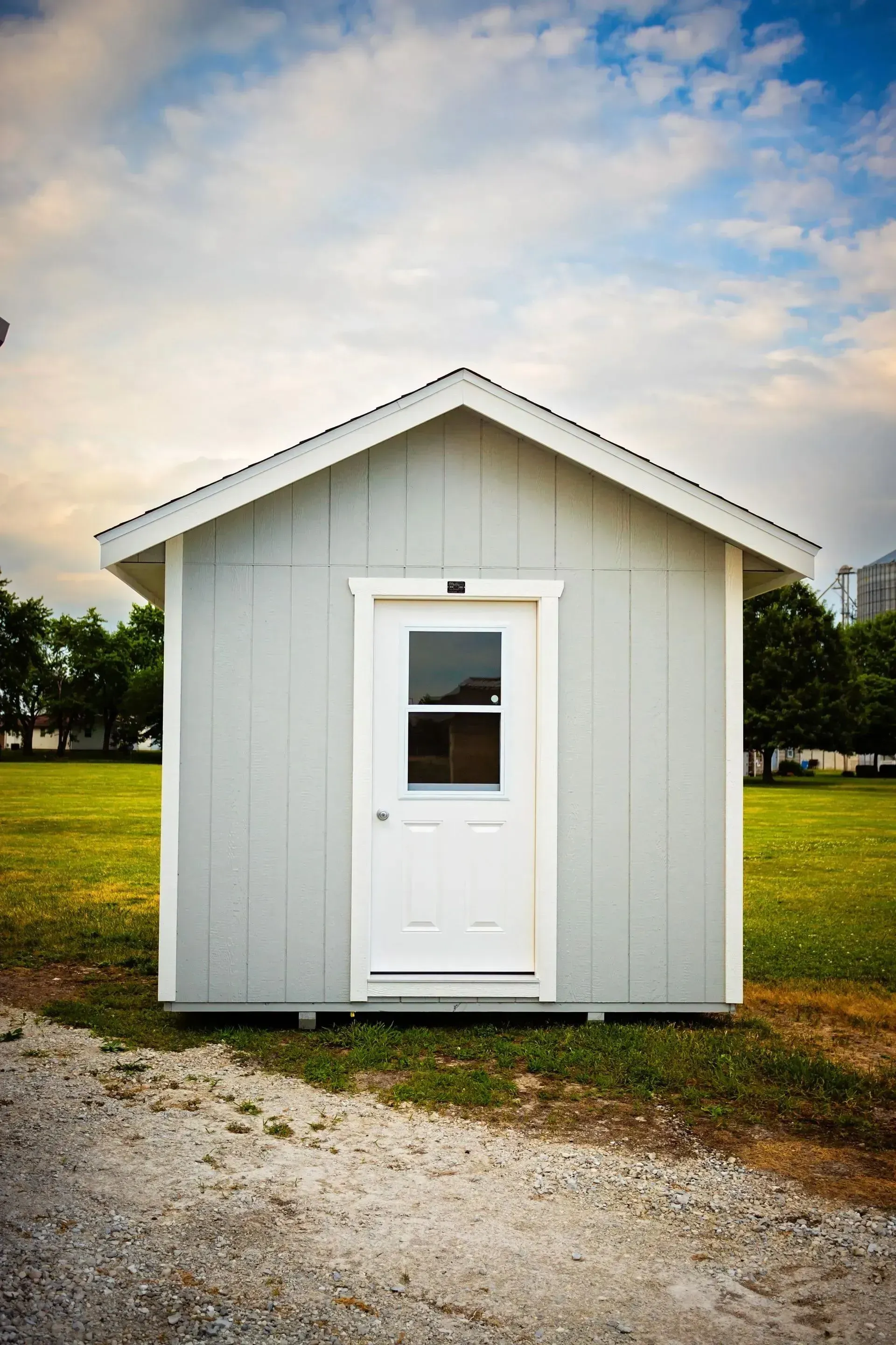 Small, gray shed with white door and trim, standing on grass, gravel in foreground.