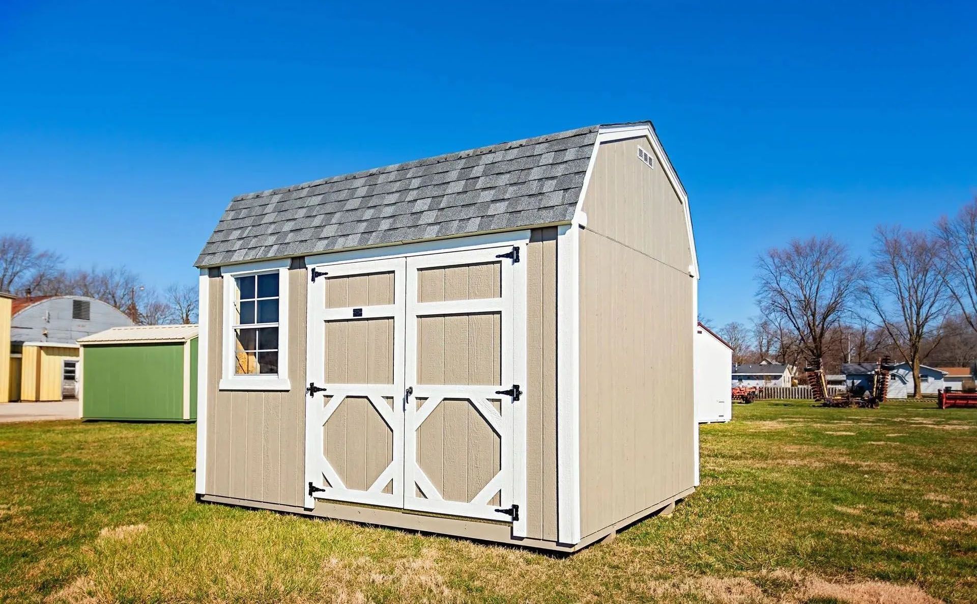 Tan barn-style shed with white trim, gray shingled roof, and blue sky background.