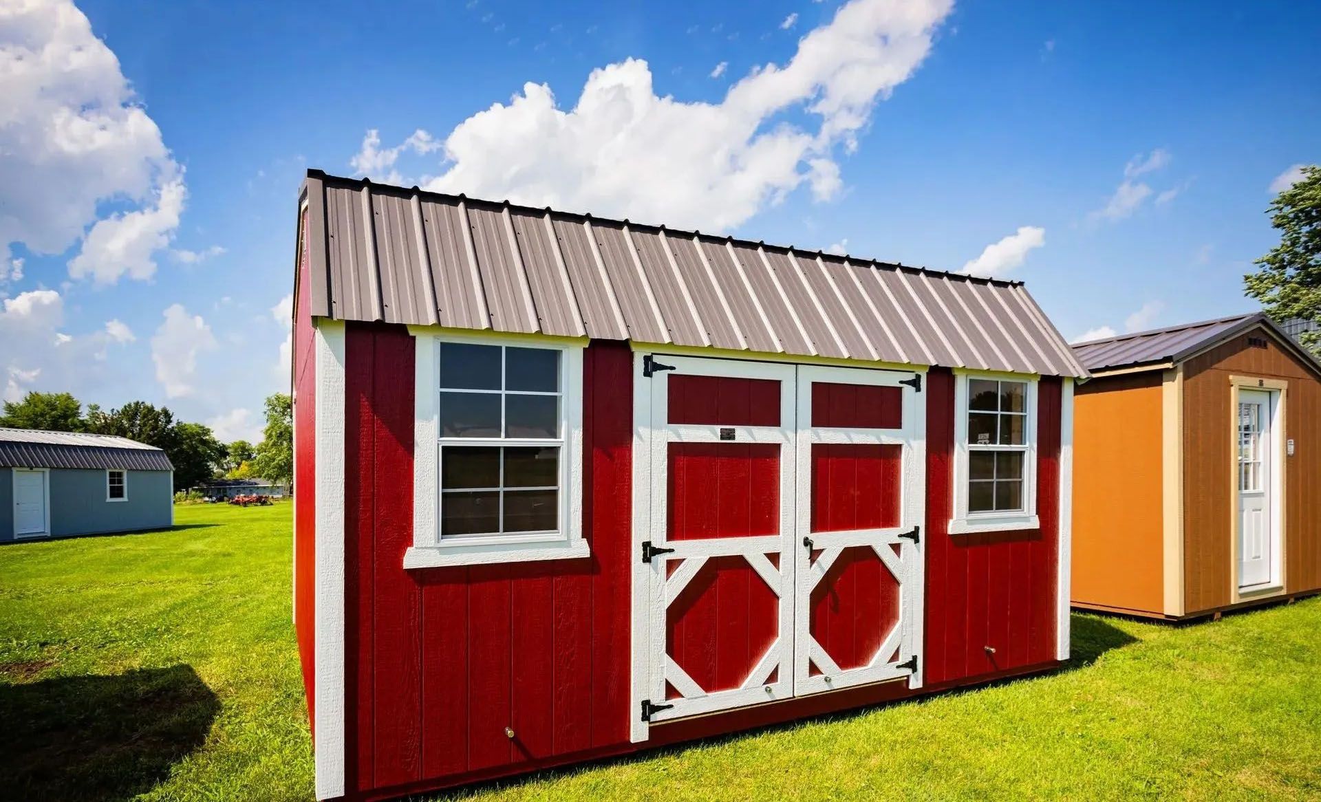 Red shed with white trim and doors, brown metal roof, set on grass under a blue sky.