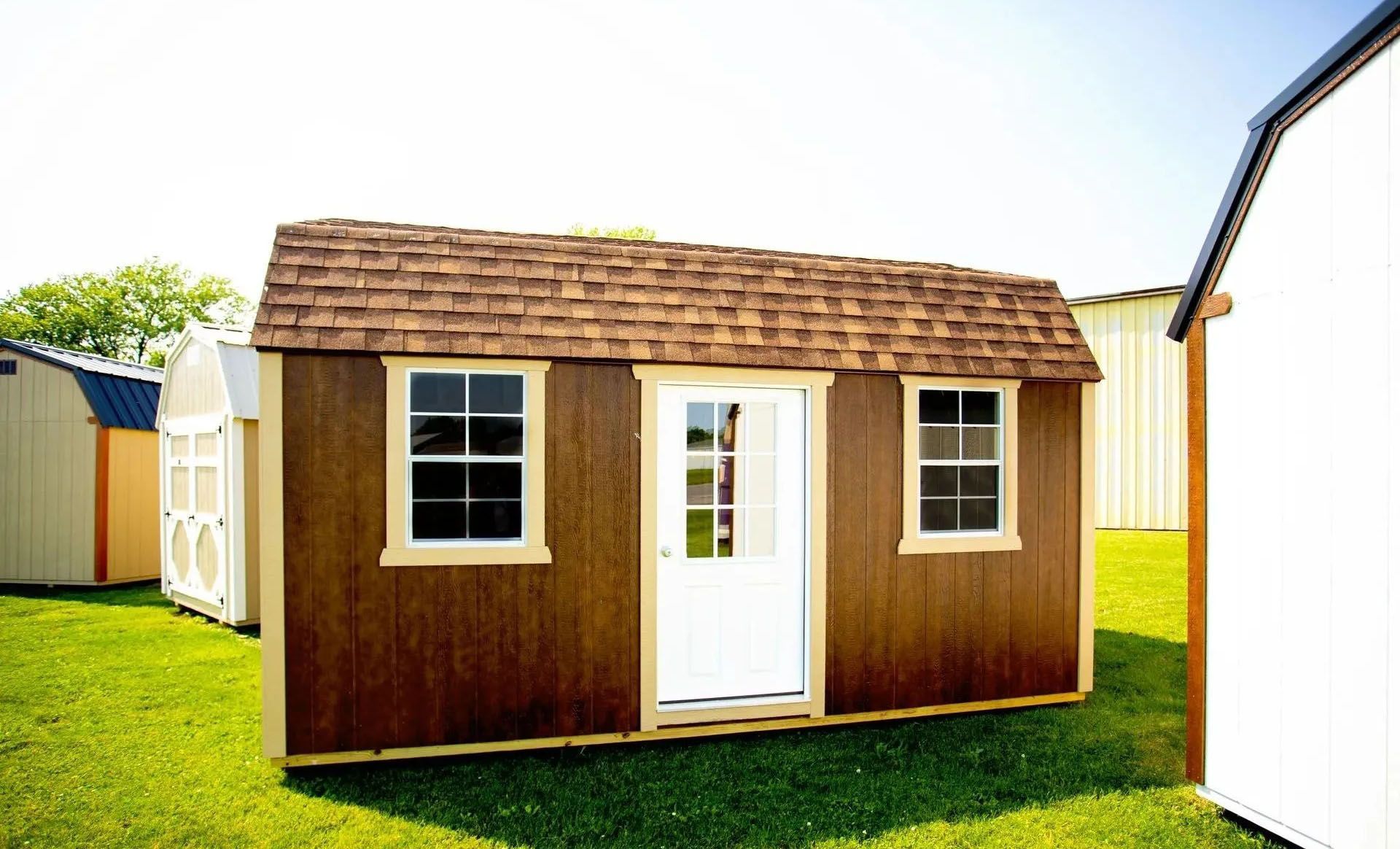 Brown shed with brown shingle roof, white trim, door, and windows, on a green lawn.