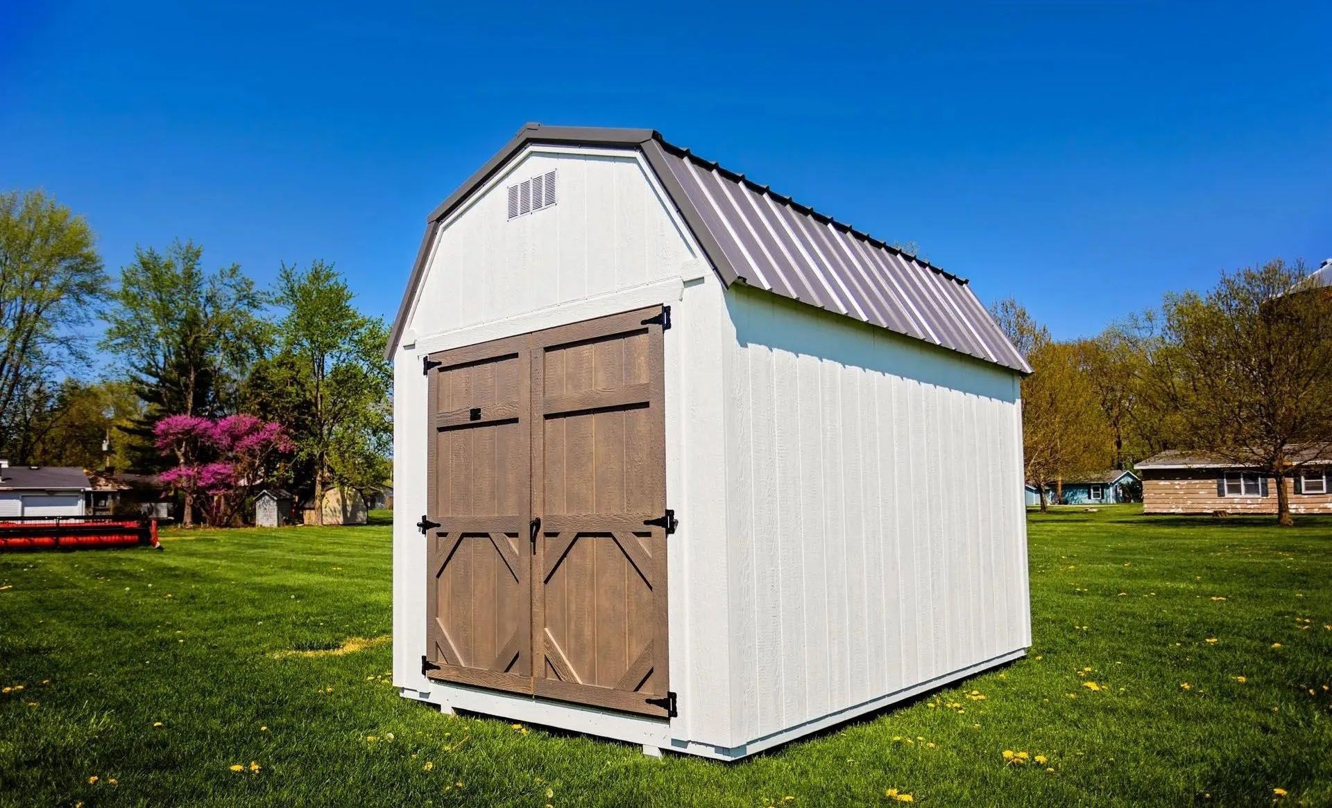 White barn-style shed with brown double doors and gray metal roof on a green lawn.