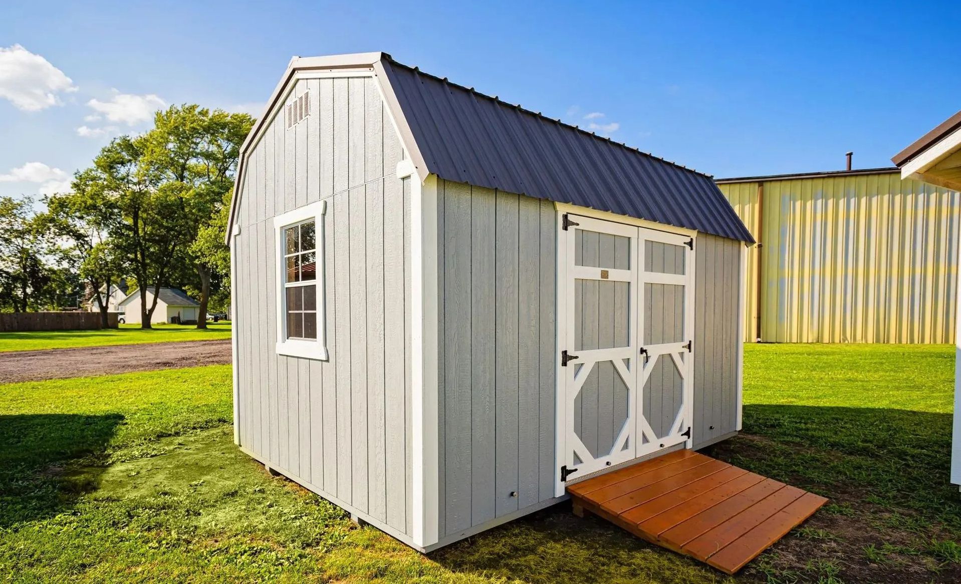 Gray shed with white trim, black roof, and brown ramp on green grass under a blue sky.