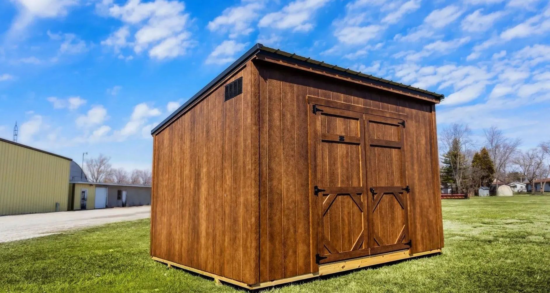 Wooden storage shed with brown siding and double doors, set on grass under a blue sky.