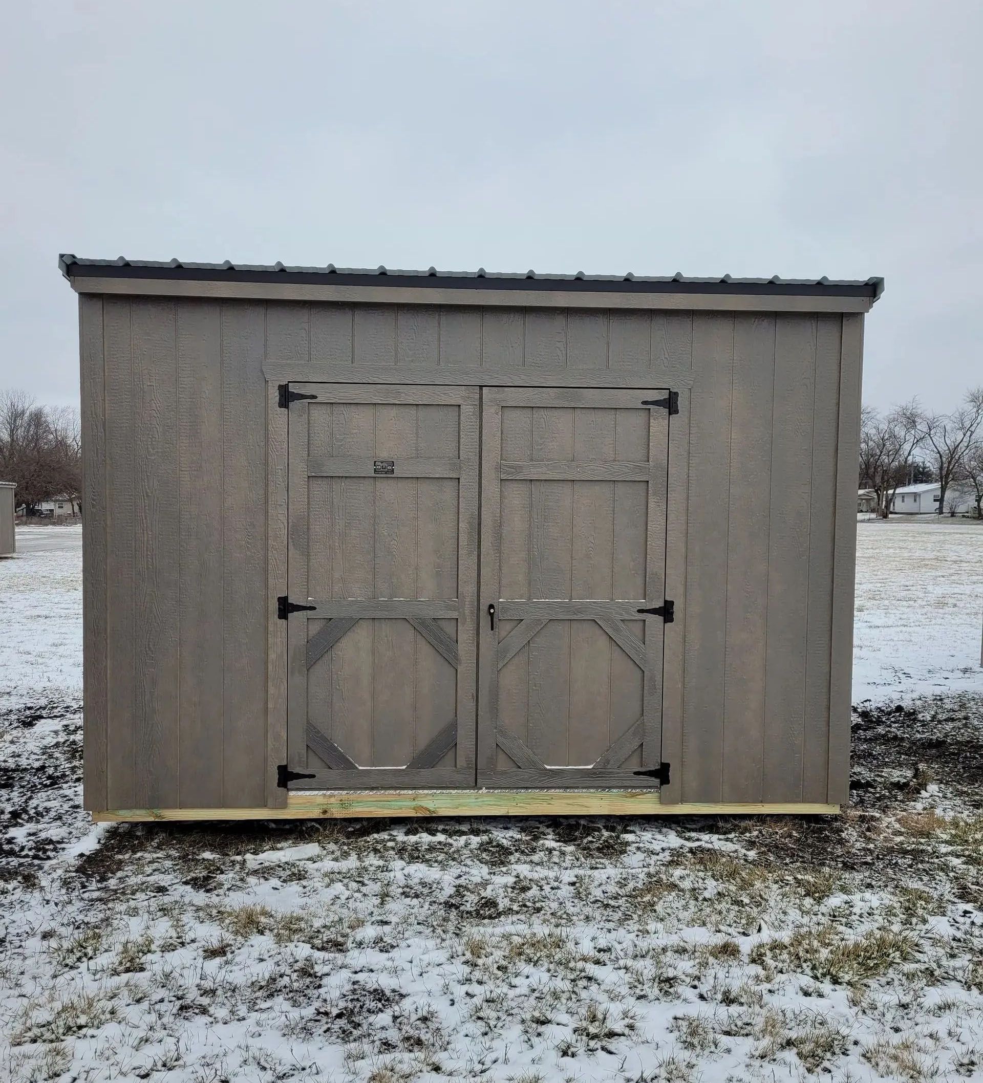 Gray wooden shed with double doors, set in a snowy field under a cloudy sky.