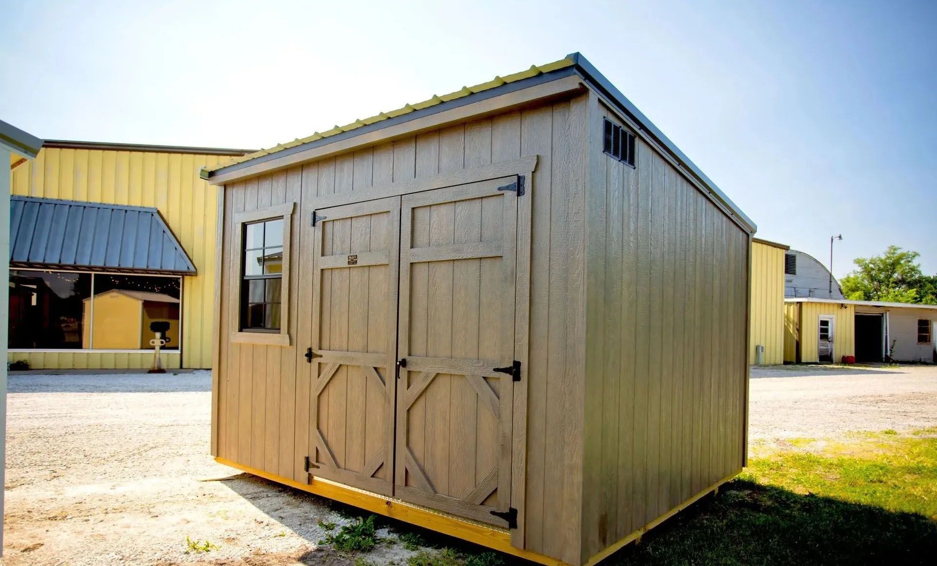 Tan shed with double doors, window, and ventilation, sitting outside on gravel and grass.