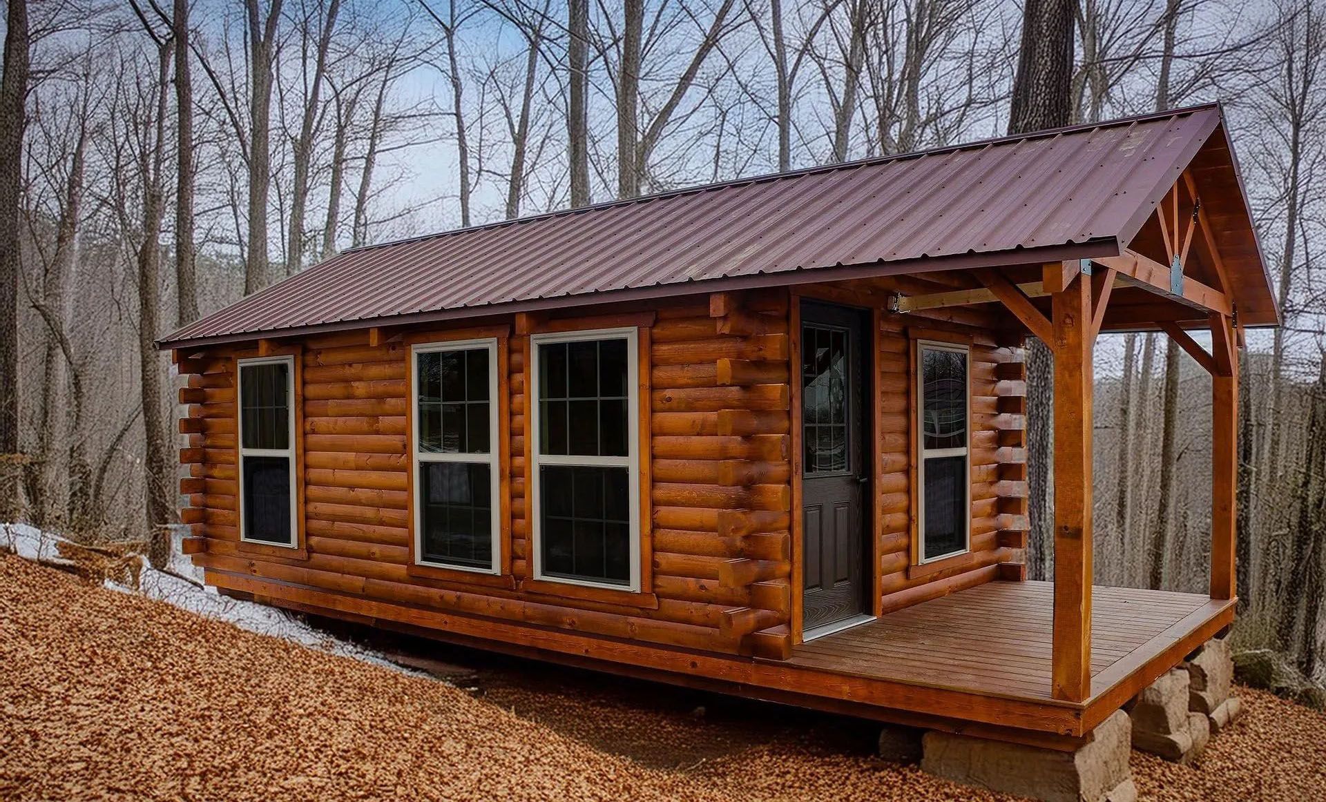 Small log cabin with porch in a wooded setting. Brown logs, dark metal roof, and windows visible.