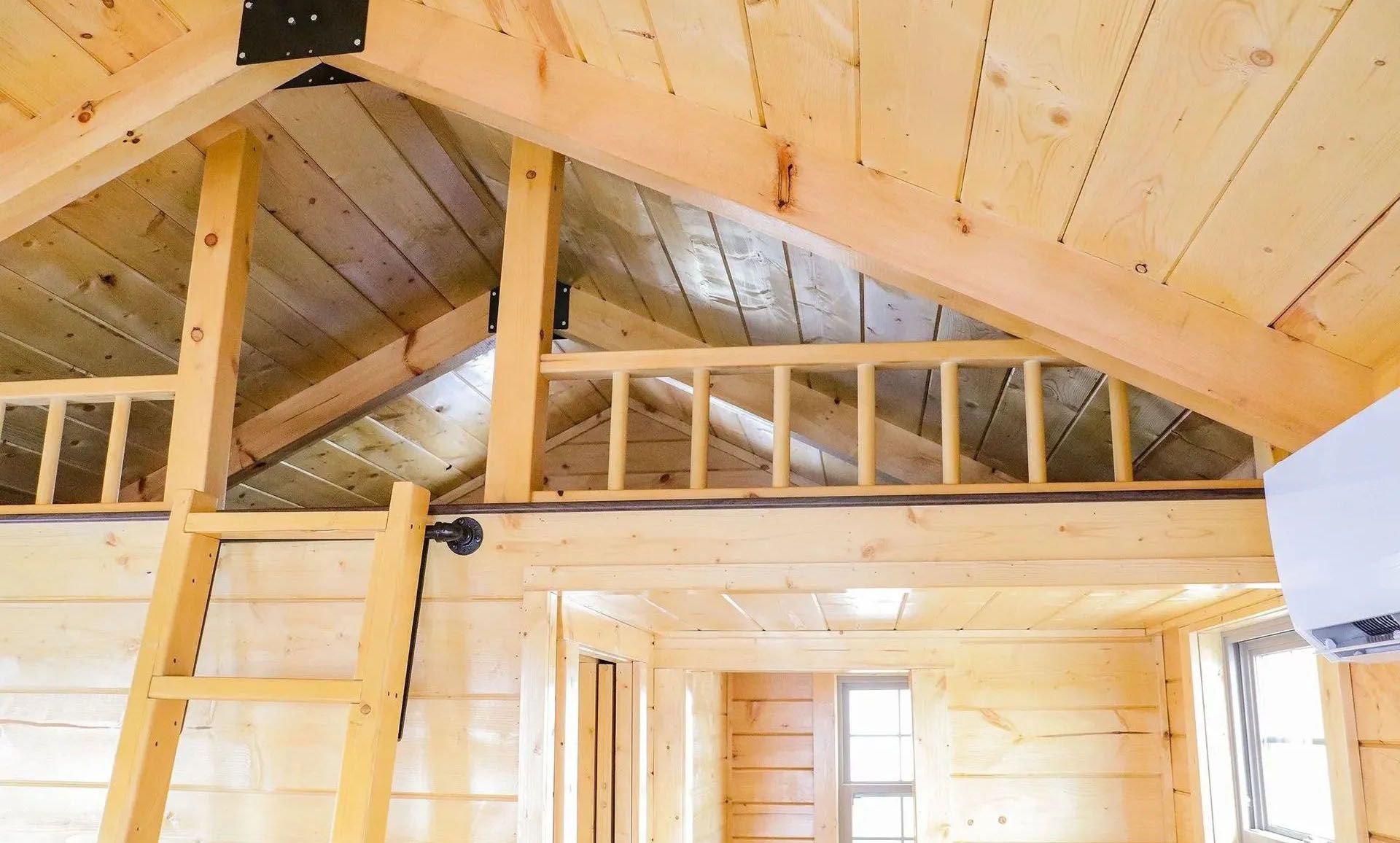 Interior of a small wooden cabin with a loft area, ladder, and a railed balcony.