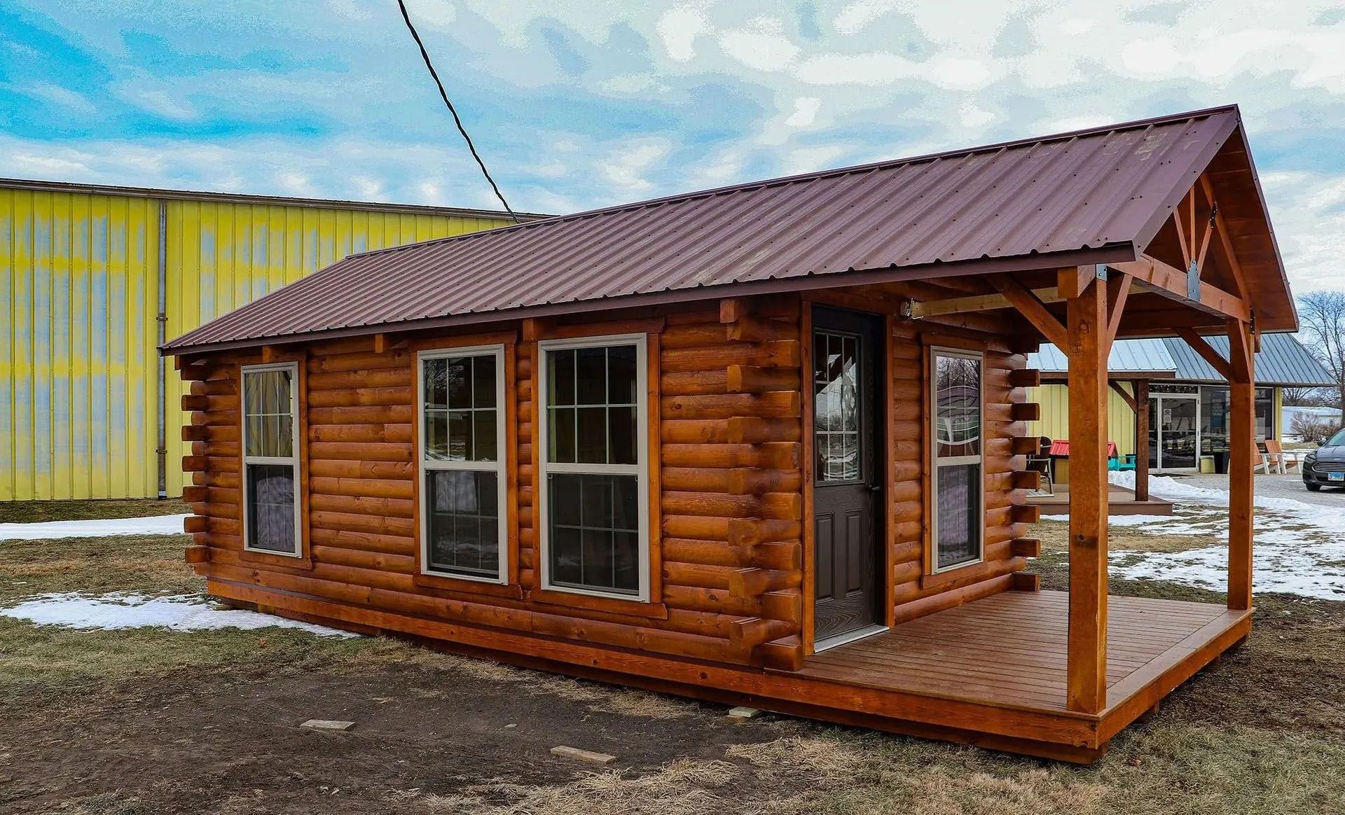 Log cabin with brown metal roof, porch, and windows, set in a snowy, grassy area.