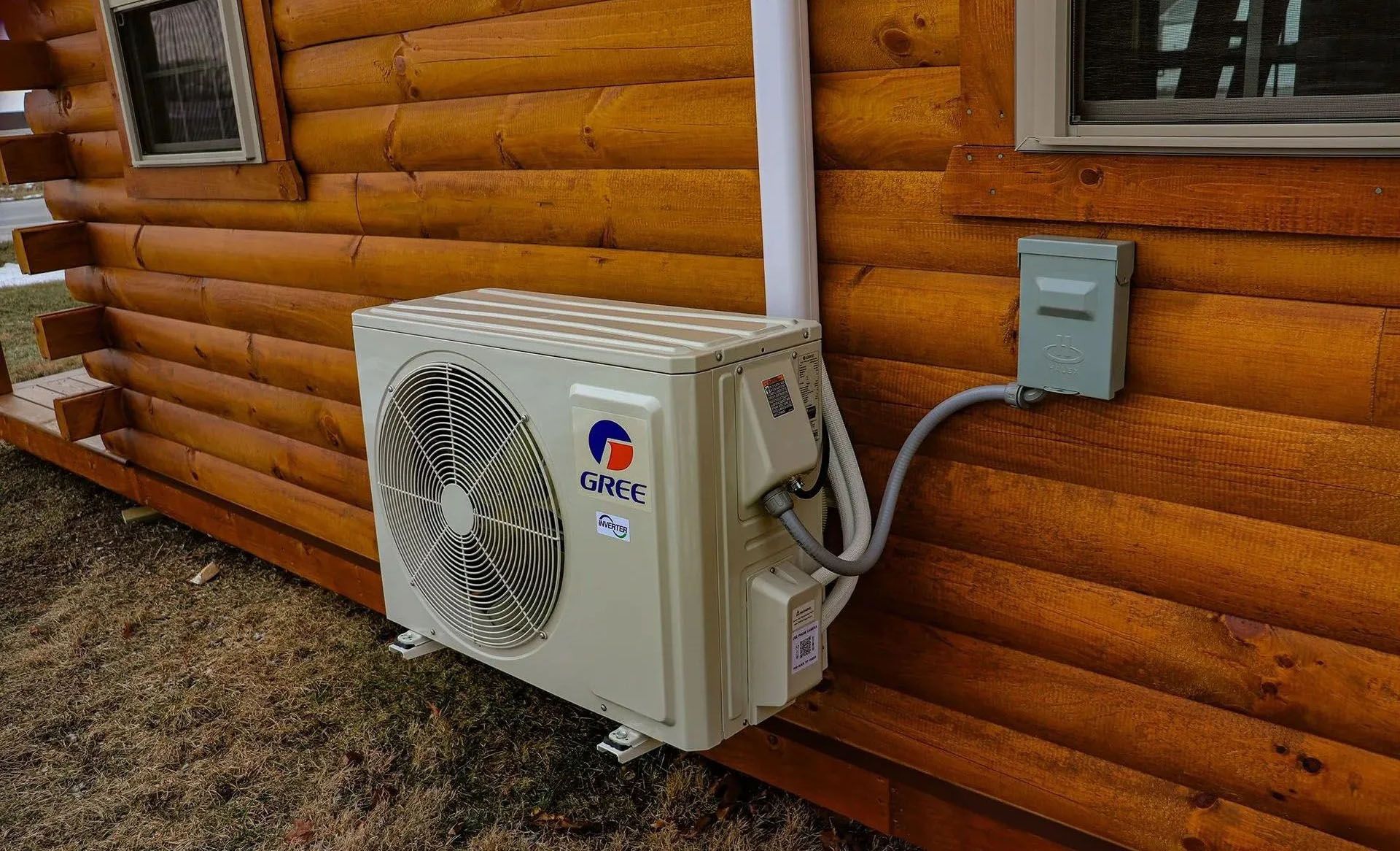 An air conditioning unit mounted on a log cabin wall with electrical conduit and a grey electrical box.