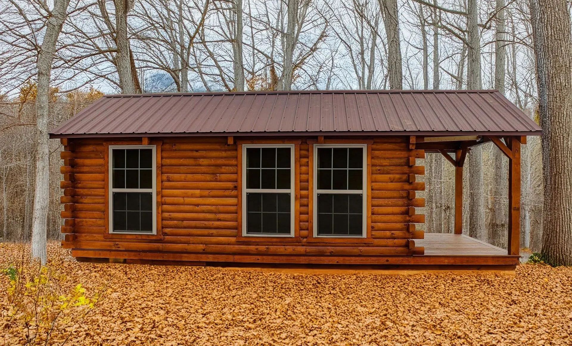 Log cabin with brown metal roof, three windows, and small porch in a forest of trees and fallen leaves.