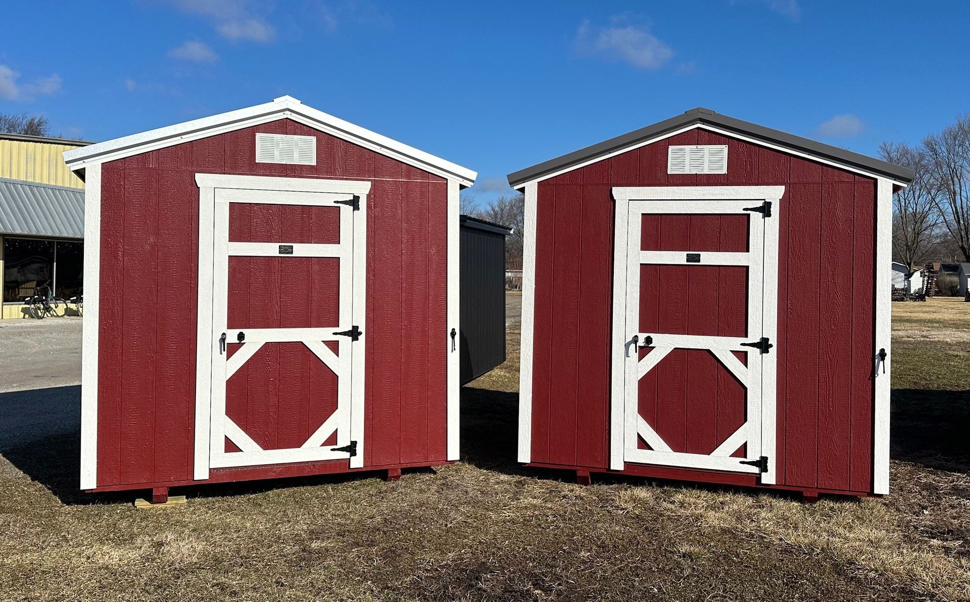 Two red sheds with white trim, doors, and roofs, standing in a grassy area under a blue sky.