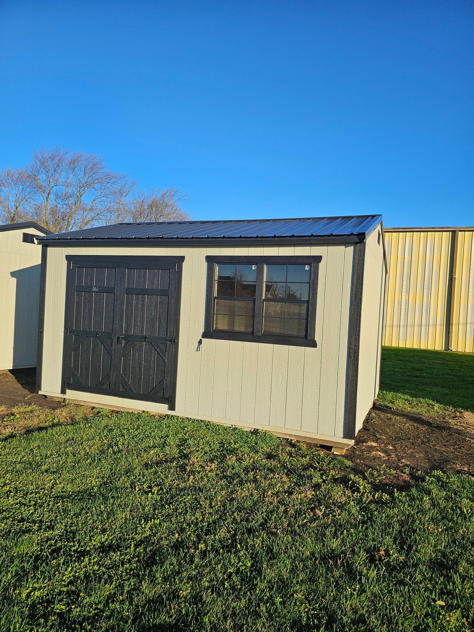 Small shed with black door, window, and roof against a blue sky, in a grassy area.