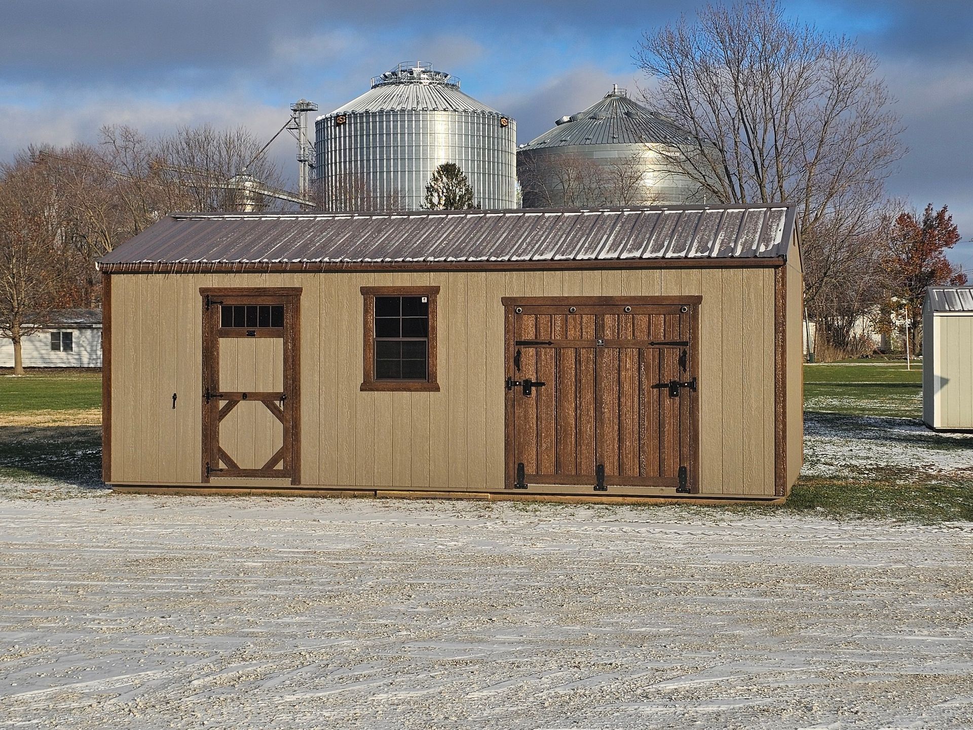 Tan shed with brown trim and metal roof, two silos in background.