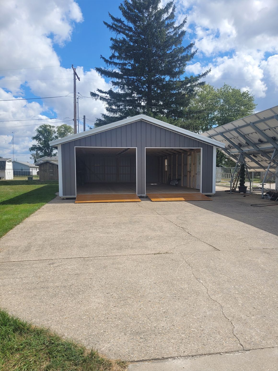 Gray detached garage with two open bays, gravel driveway, and a large evergreen tree under a blue sky.