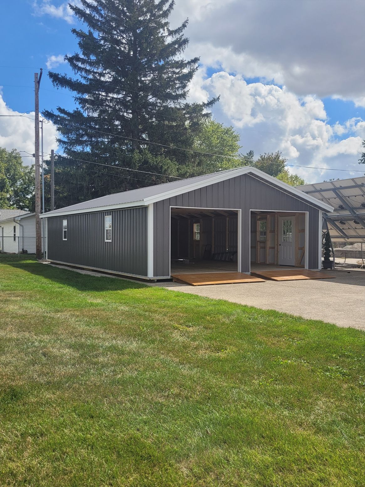 Gray metal garage with two open bays beside a grassy yard and a large tree under a partly cloudy sky.