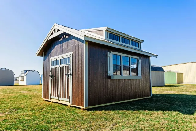 Brown shed with a light-colored trim and a small window above the larger window, set on a green grassy field.