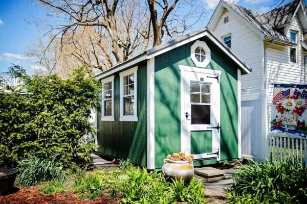 Green shed with white trim, door, and windows in a yard, with flowers and a white house.