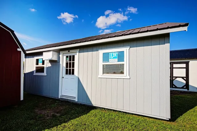 Gray storage shed with metal roof and white trim, set on green grass under a blue sky.