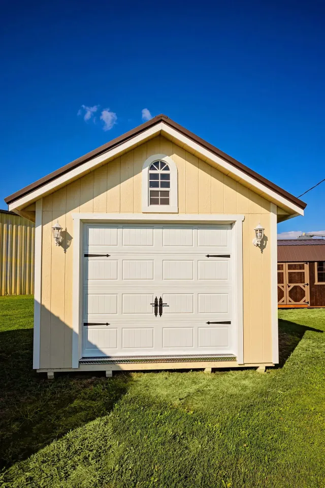 Yellow shed with white garage door and small arched window; set in green grass against a blue sky.