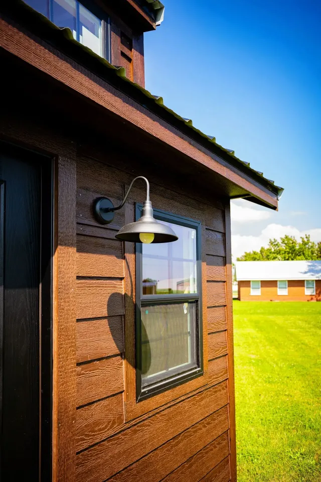 Brown-sided tiny house exterior with window, lamp, and green lawn in bright sunlight.