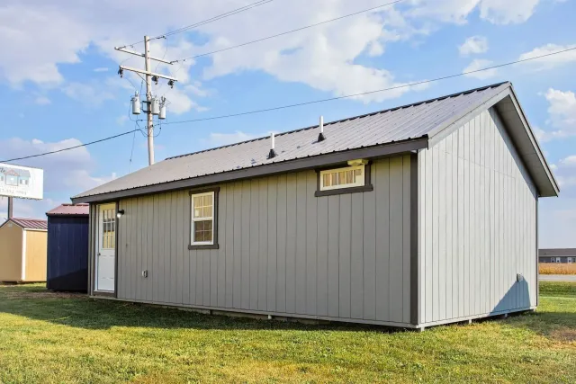 Gray shed with metal roof, windows, and door, set on a grassy field under a cloudy sky.