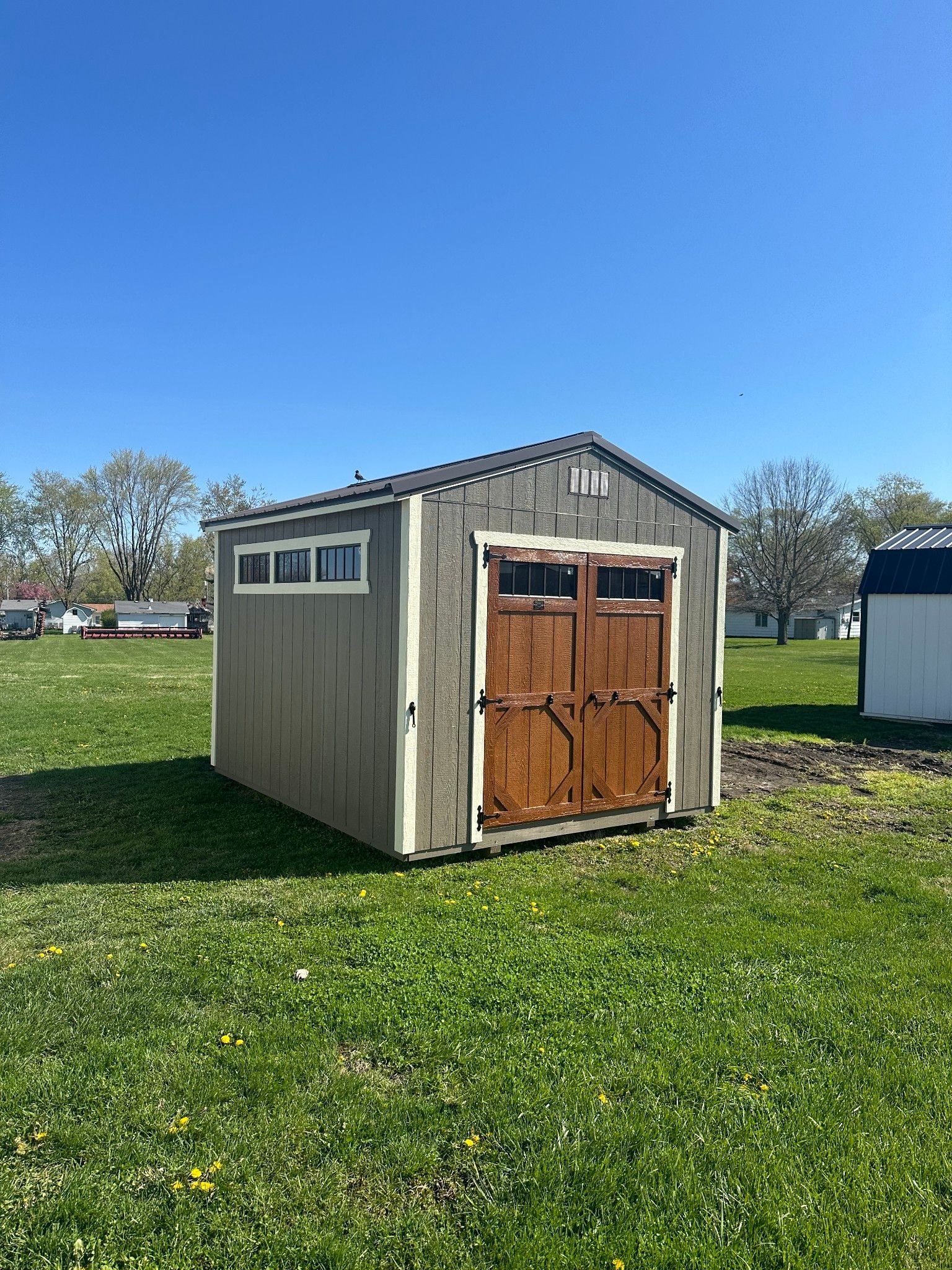 Gray storage shed with brown double doors and small windows on a grassy lawn under a blue sky.
