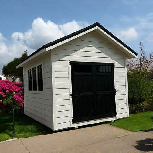 White shed with black doors, windows, and roof on green grass next to a paved path and pink flowers.