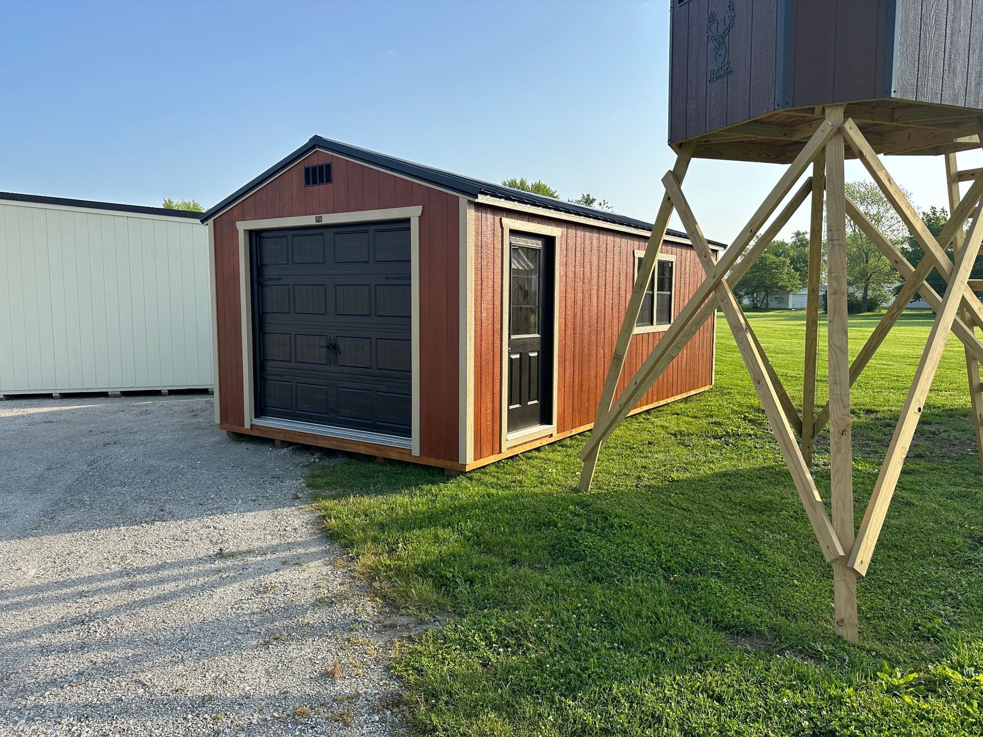 A red wooden shed with a black garage door and side door sits on a gravel lot next to a tall wooden deer stand.