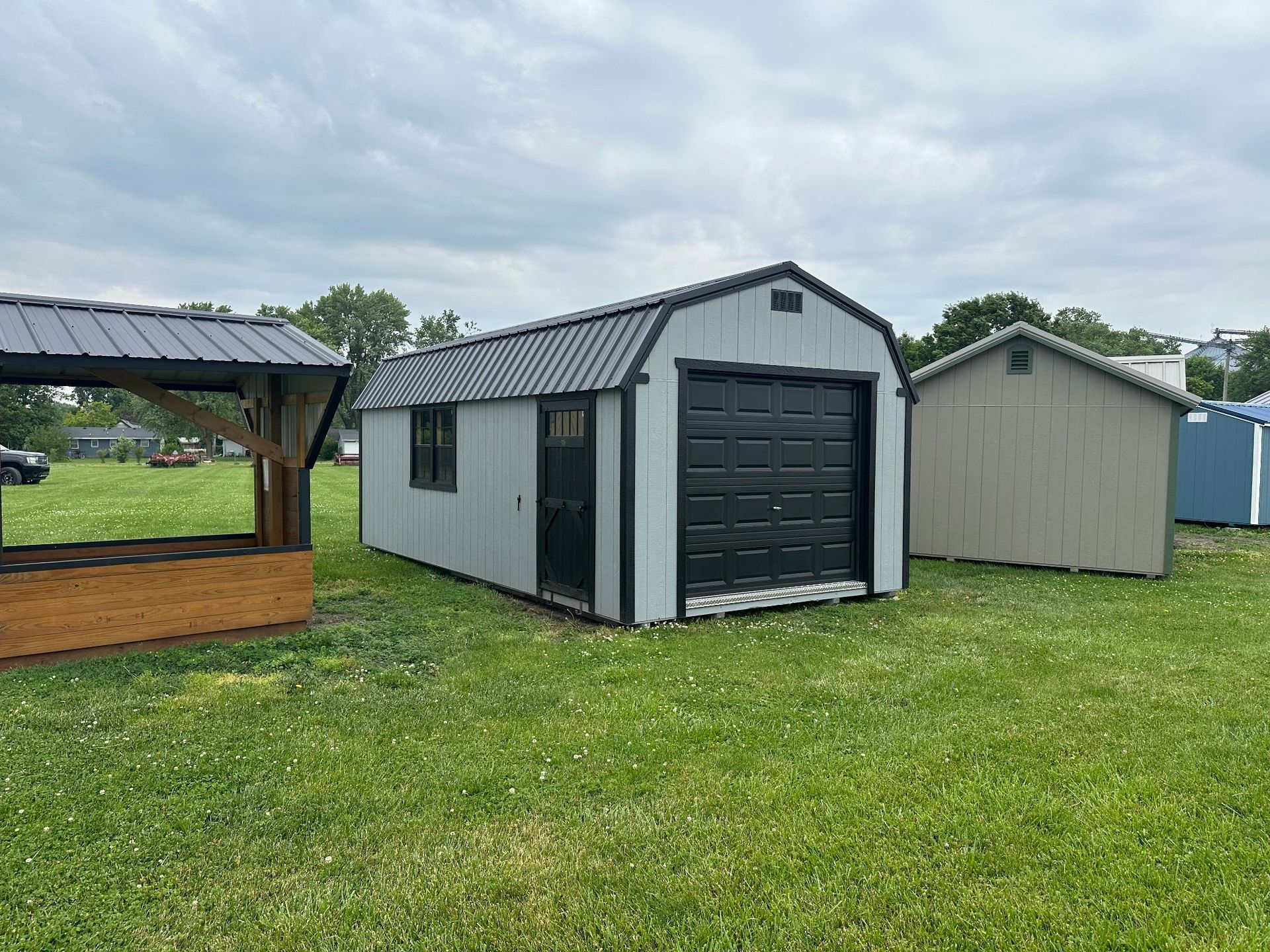 Three outdoor storage structures, including a grey shed with a black garage door, sit on a grassy field under a cloudy sky.