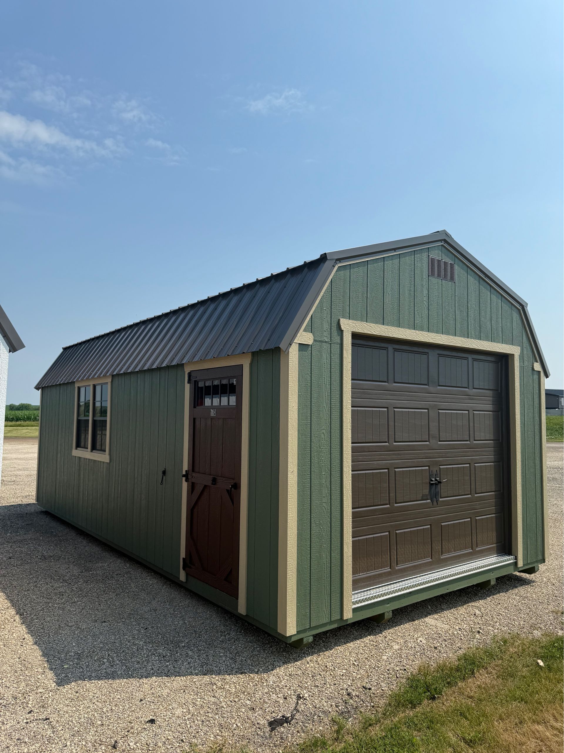 A green wooden shed with a dark metal roof, a brown side door, and a brown roll-up garage door on a gravel lot.