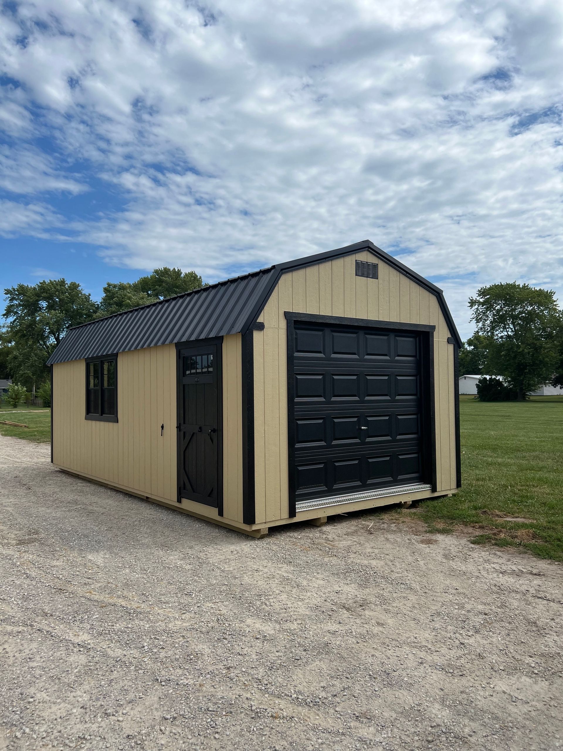 A beige shed with a black roof, door, and garage door sitting on a gravel lot under a cloudy sky.