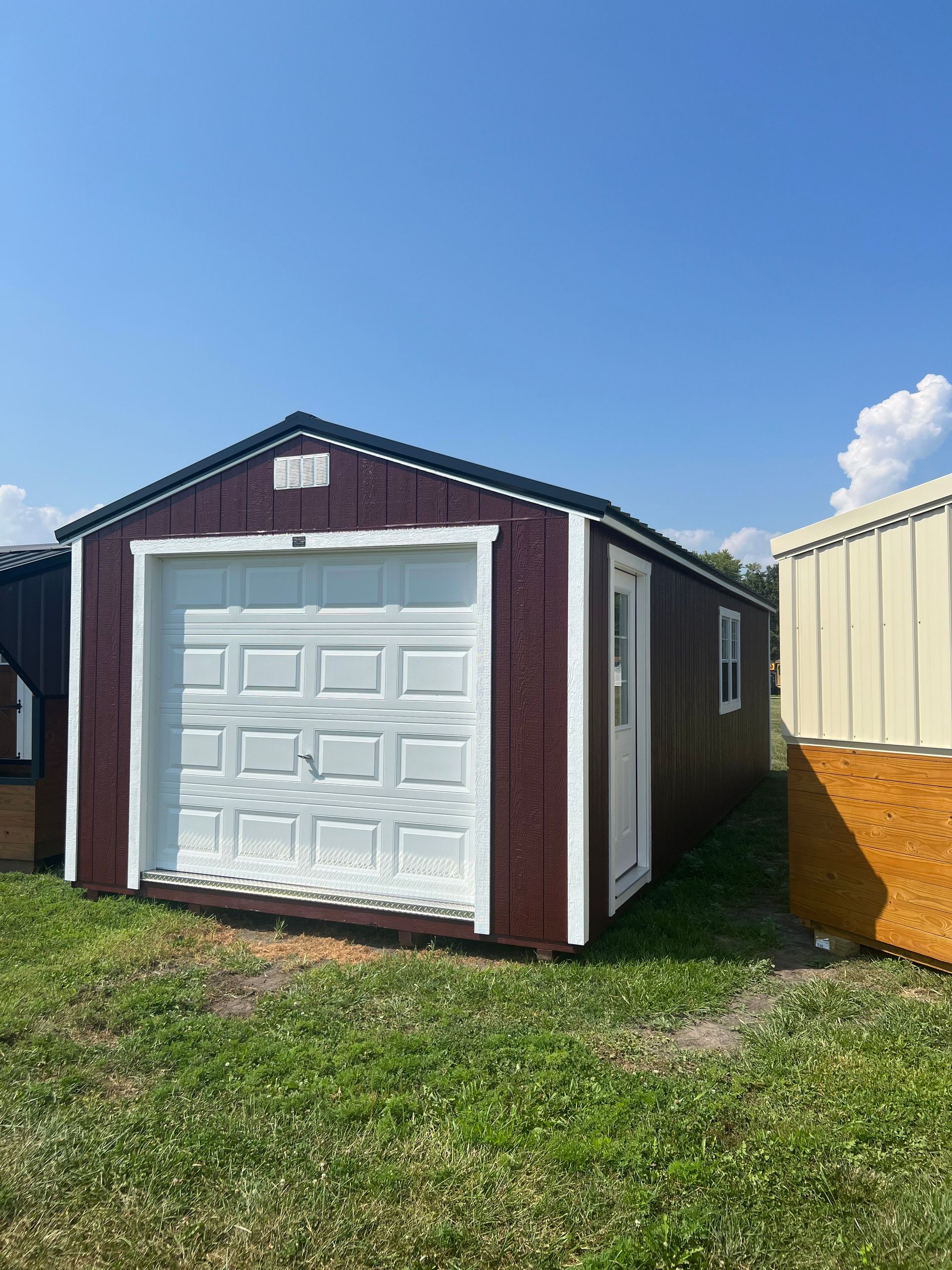 A burgundy storage shed with a white garage door and side door, positioned on grass under a clear blue sky.