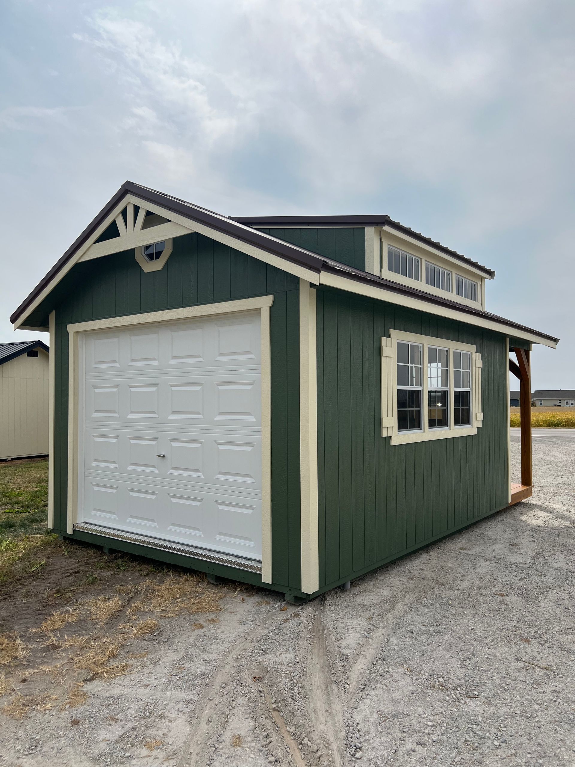 A green, single-car wooden garage with a white roll-up door, side windows, and a dormer roof on a gravel lot.