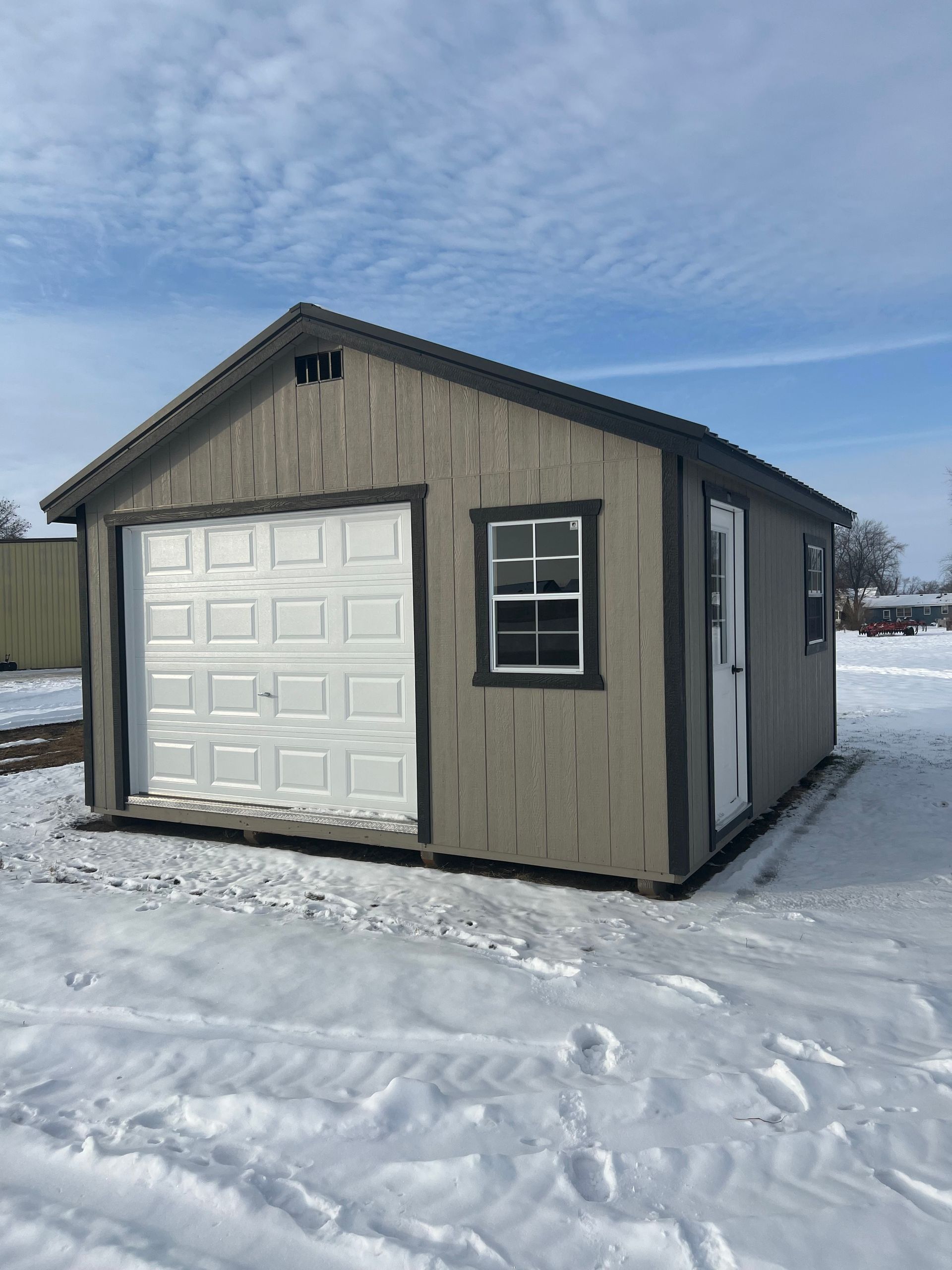 A tan, gable-roofed shed with a white garage door, a window, and a side entry door, sitting in a snow-covered field.