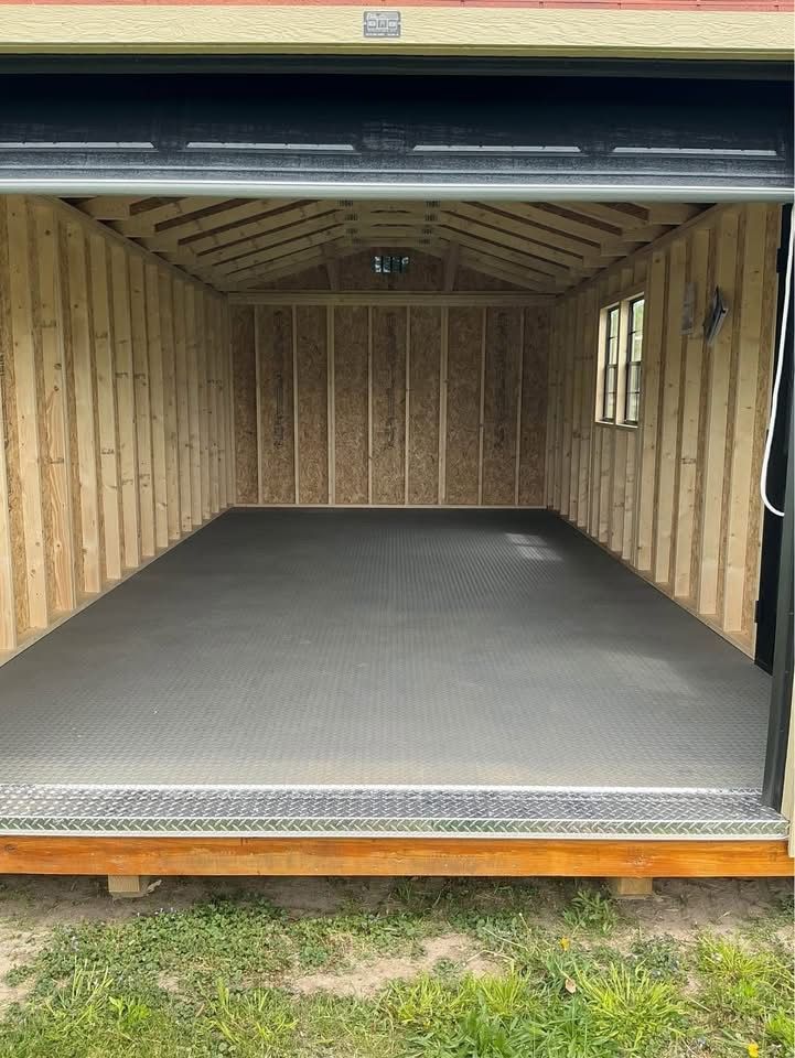 An open shed with light-colored wooden walls and roof rafters, featuring a gray, diamond-plate rubber floor mat.